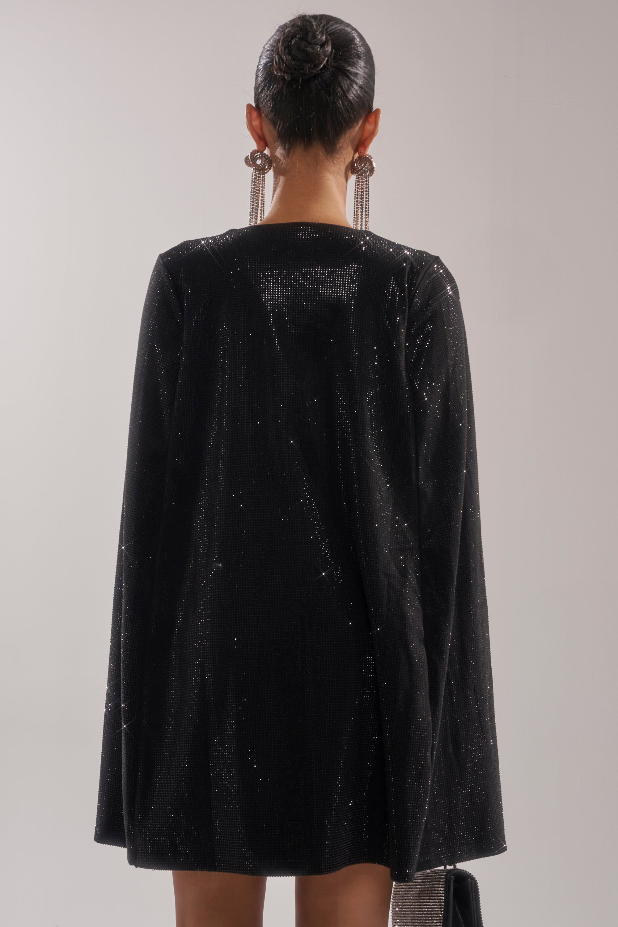 A person with dark hair in a sleek bun is seen from behind wearing the LETS DANCE RHINESTONE MINI DRESS and long, dangling earrings against a plain background.