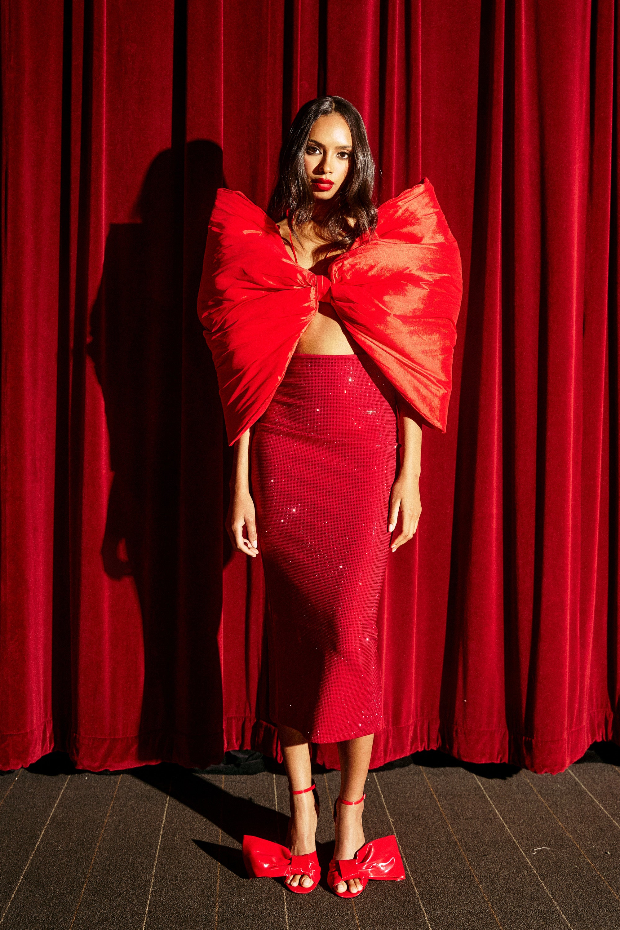A woman poses before red velvet curtains in the glamorous ICED OUT RHINESTONE MAXI SKIRT and a dramatic bow top. Her long dark hair and red heels with bow accents finish the dazzling, monochromatic look.