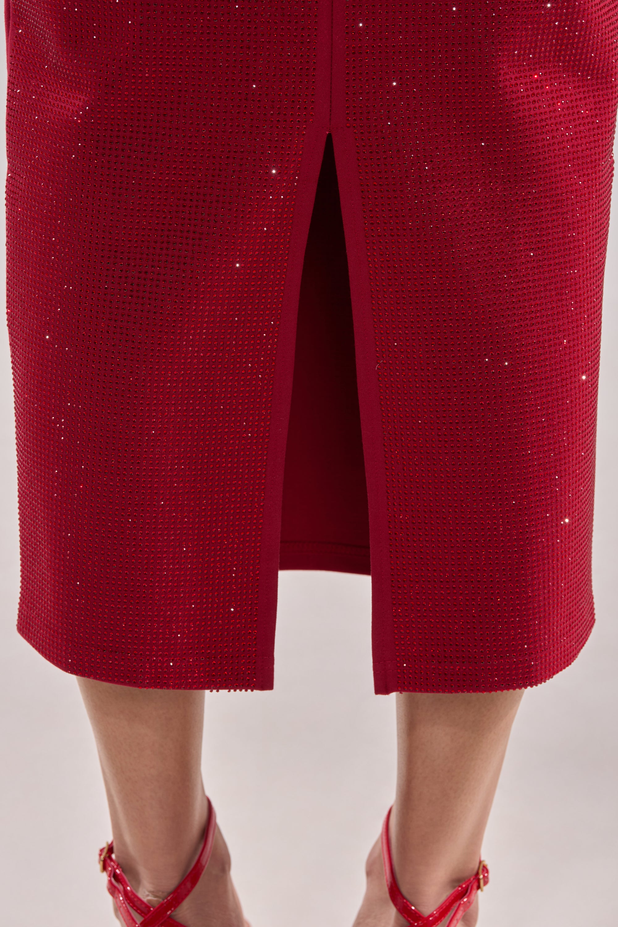 Cropped view of a person wearing the ICED OUT RHINESTONE MAXI SKIRT with a front slit, styled with red high-heeled sandals. The sparkling red rhinestone design stands out against a blurred, neutral background.
