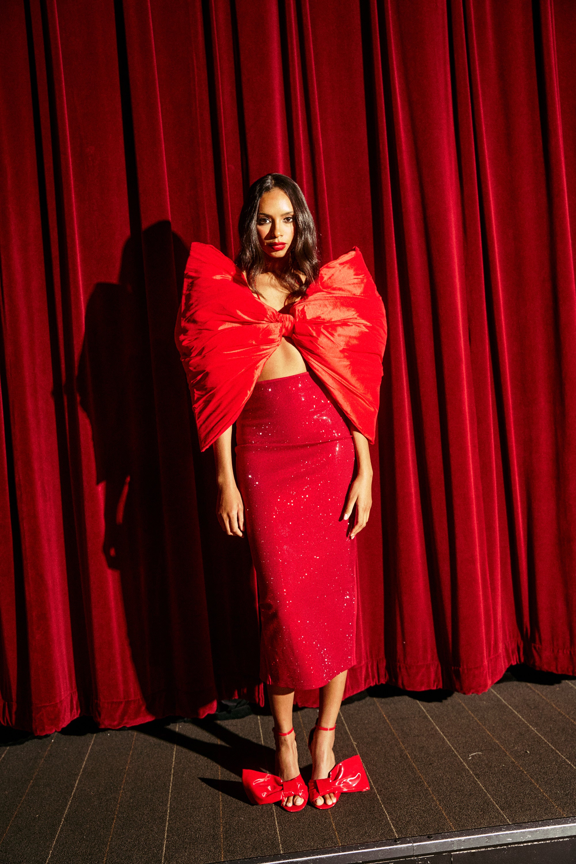 A woman with long hair and a confident look stands before red velvet curtains wearing an ICED OUT RHINESTONE MAXI SKIRT, red bow heels, and a large red bow-shaped top covering her chest and shoulders.