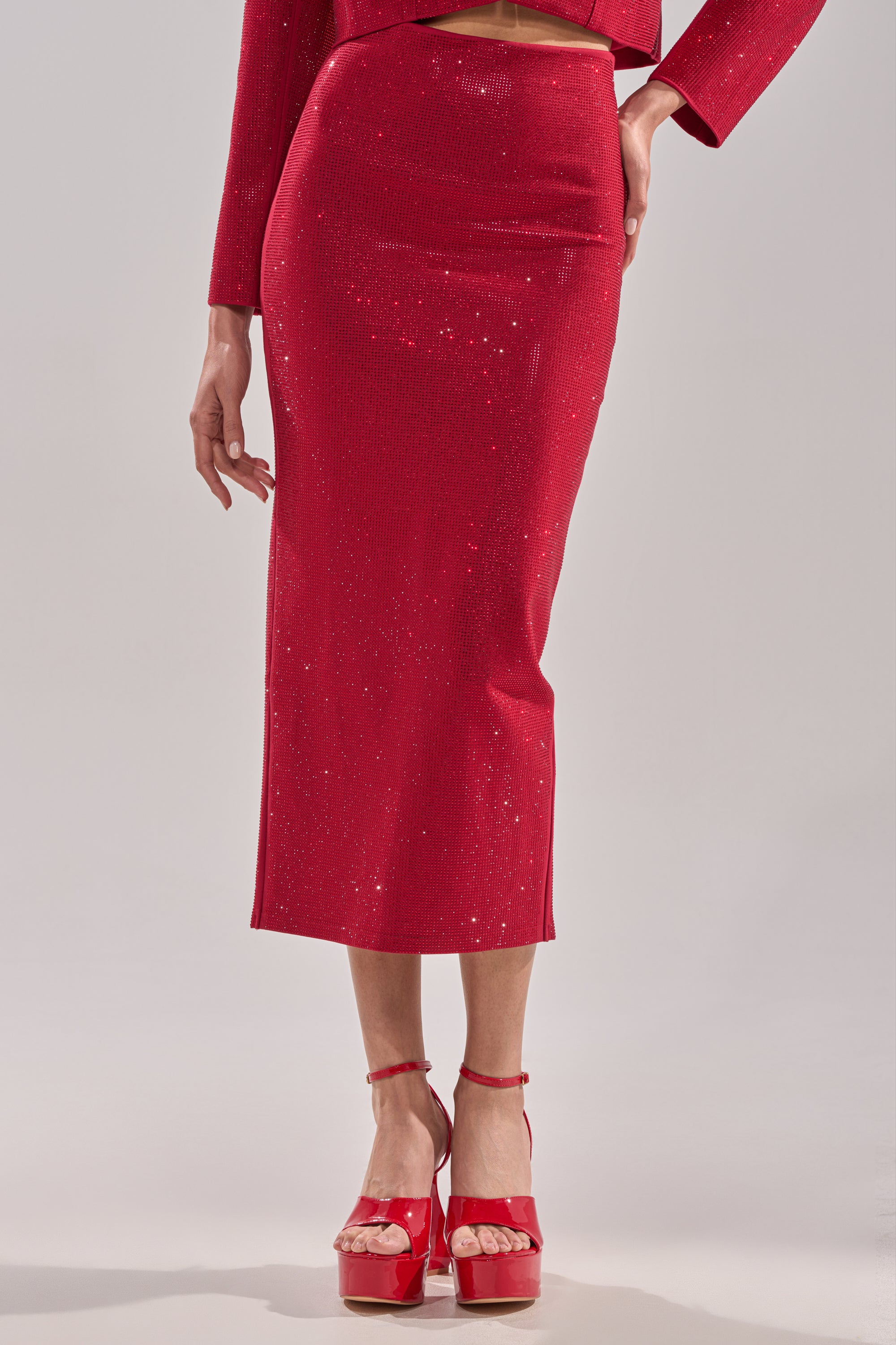 Someone wears the ICED OUT RHINESTONE MAXI SKIRT and a matching red long-sleeve top, paired with shiny red platform heels with ankle straps, standing against a plain light background.