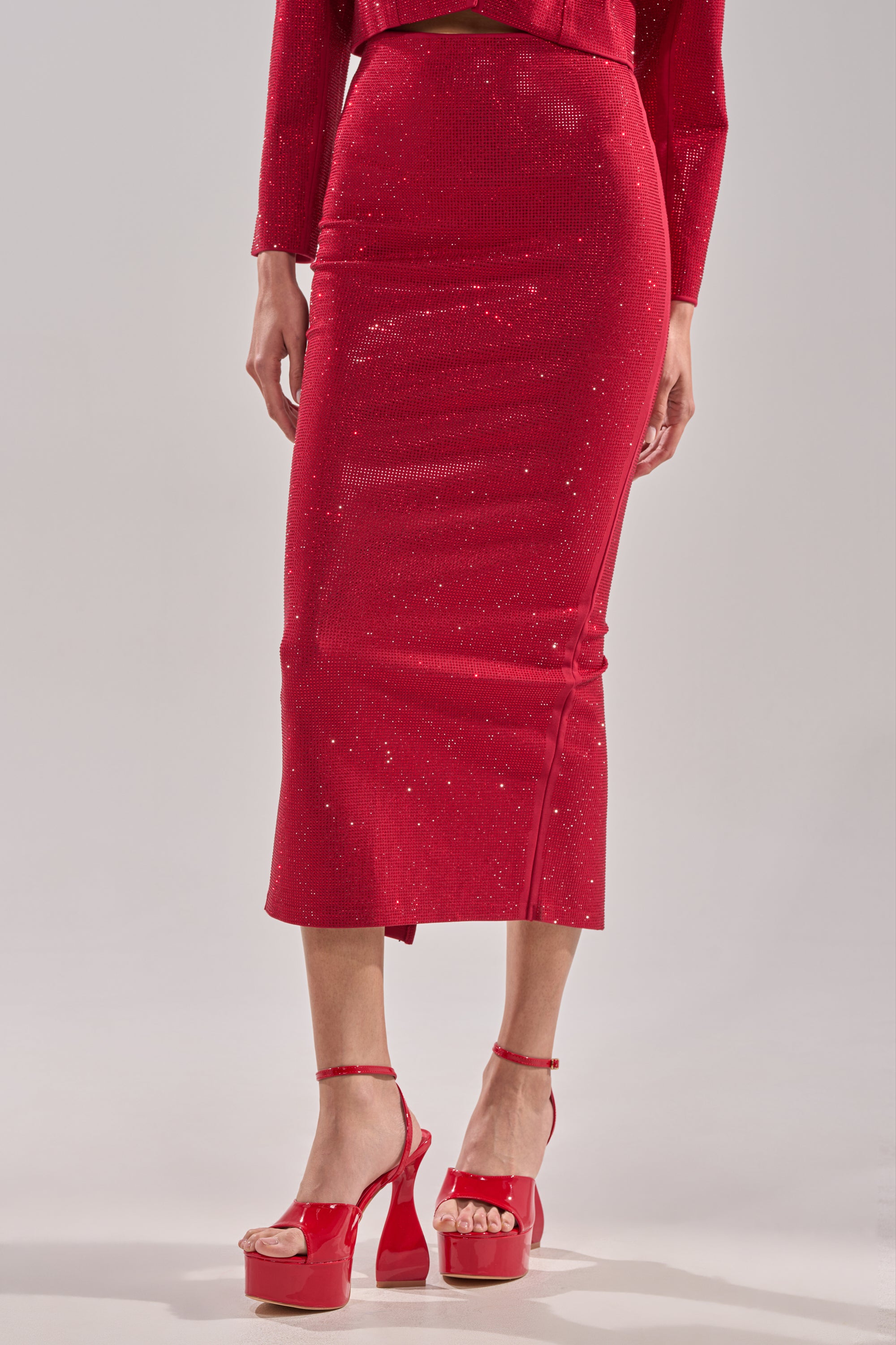 A person wears the fitted, sparkling red ICED OUT RHINESTONE MAXI SKIRT, a matching long-sleeve top, and red high-heeled platform sandals, standing against a plain light background.