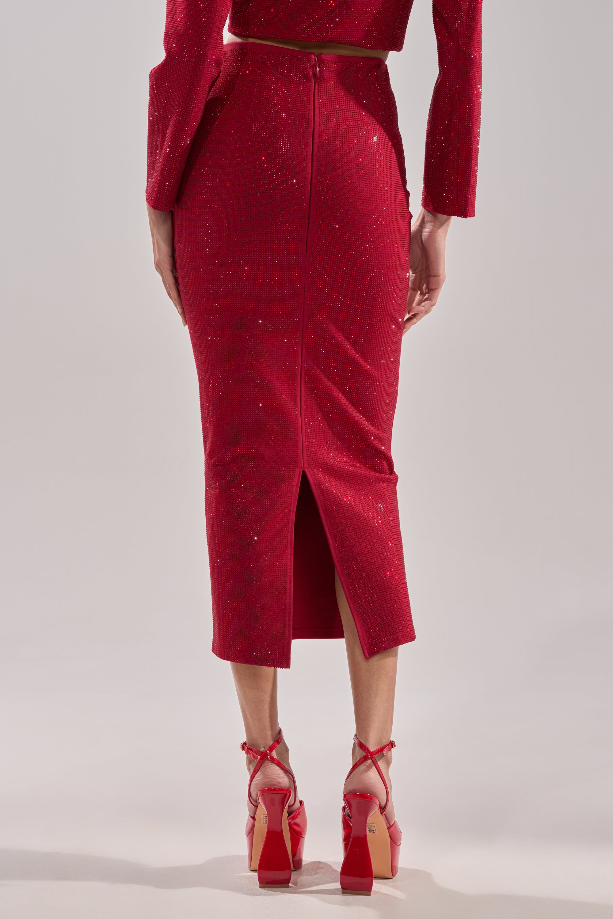 A person wearing a sparkly red ICED OUT RHINESTONE MAXI SKIRT with a back slit, a matching red top, and red high-heeled shoes with ankle straps, standing against a plain background.