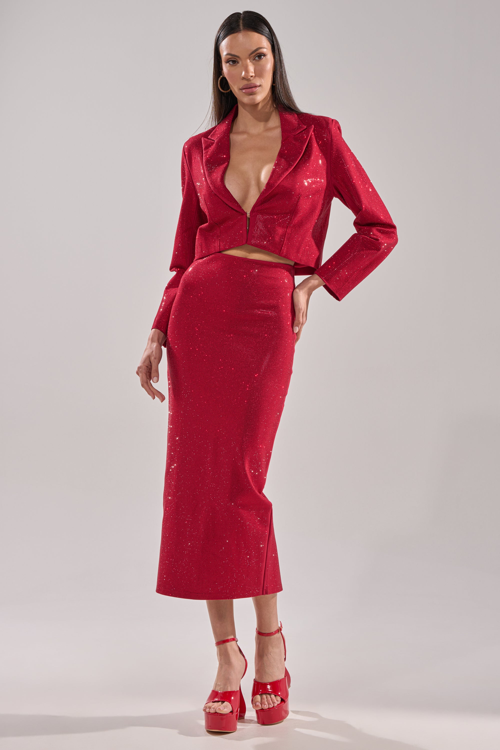 A woman models the ICED OUT RHINESTONE MAXI SKIRT and a sparkling red cropped blazer with a deep V-neck, paired with red platform heels and hoop earrings, posing against a plain background.