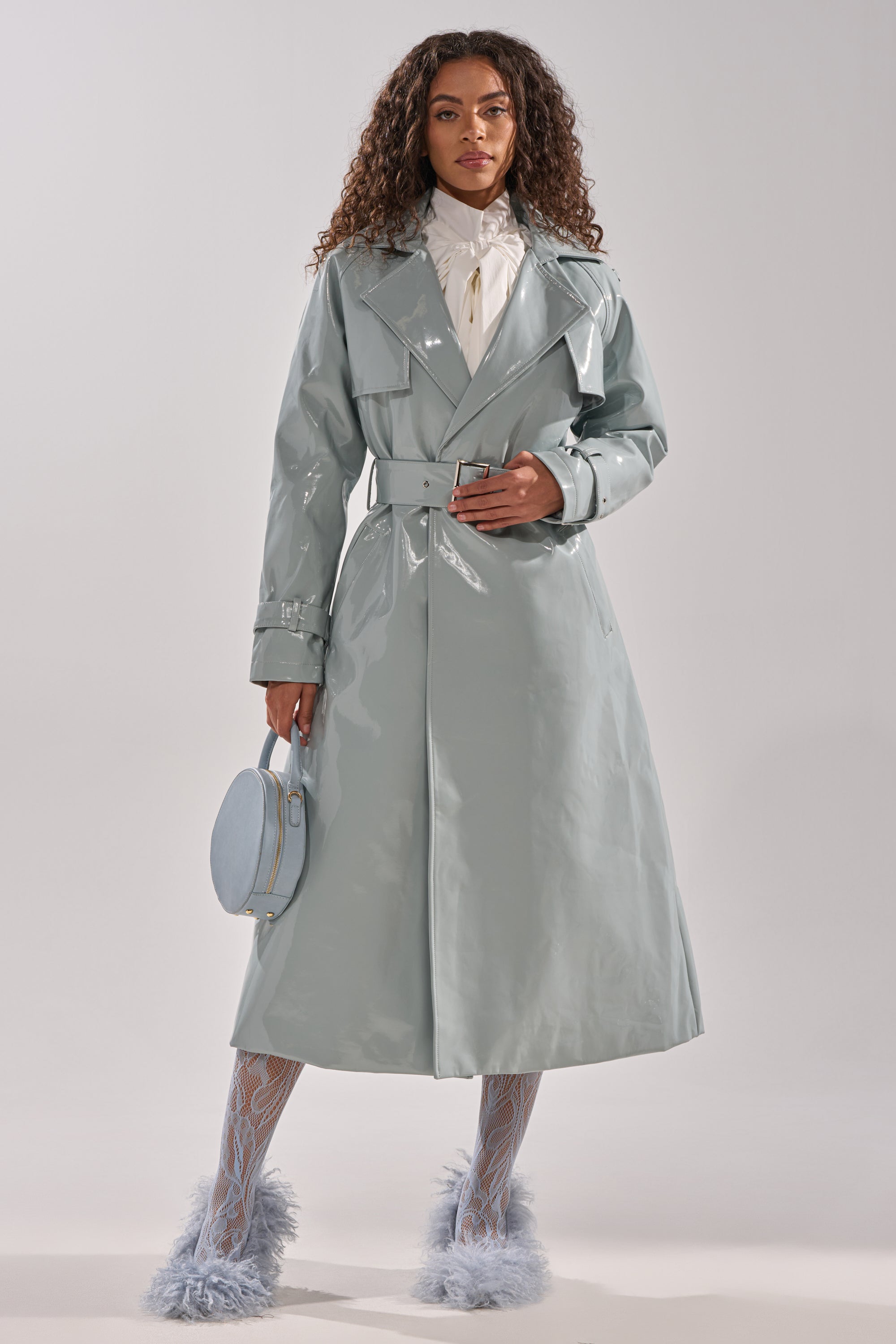 A woman with curly hair wears the LOCKED AND LOADED PATENT TRENCH in shiny light grey, styled with a white blouse, patterned tights, fluffy light blue heels, and a round light blue handbag against a plain background. 0