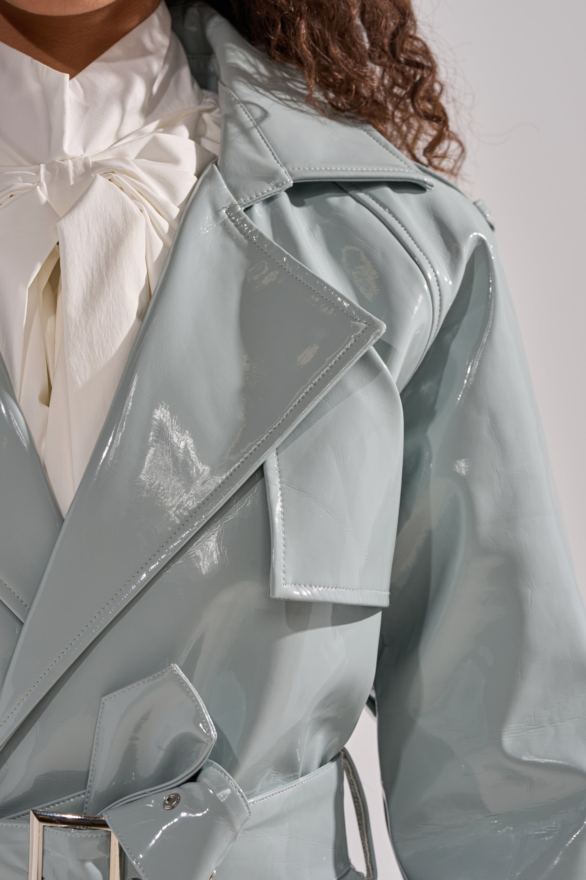 A close-up shows a person in the LOCKED AND LOADED PATENT TRENCH, a shiny light blue vegan patent leather coat, worn over a white blouse with a large neck bow. Only the shoulders, upper torso, and part of the face and hair are visible. 6