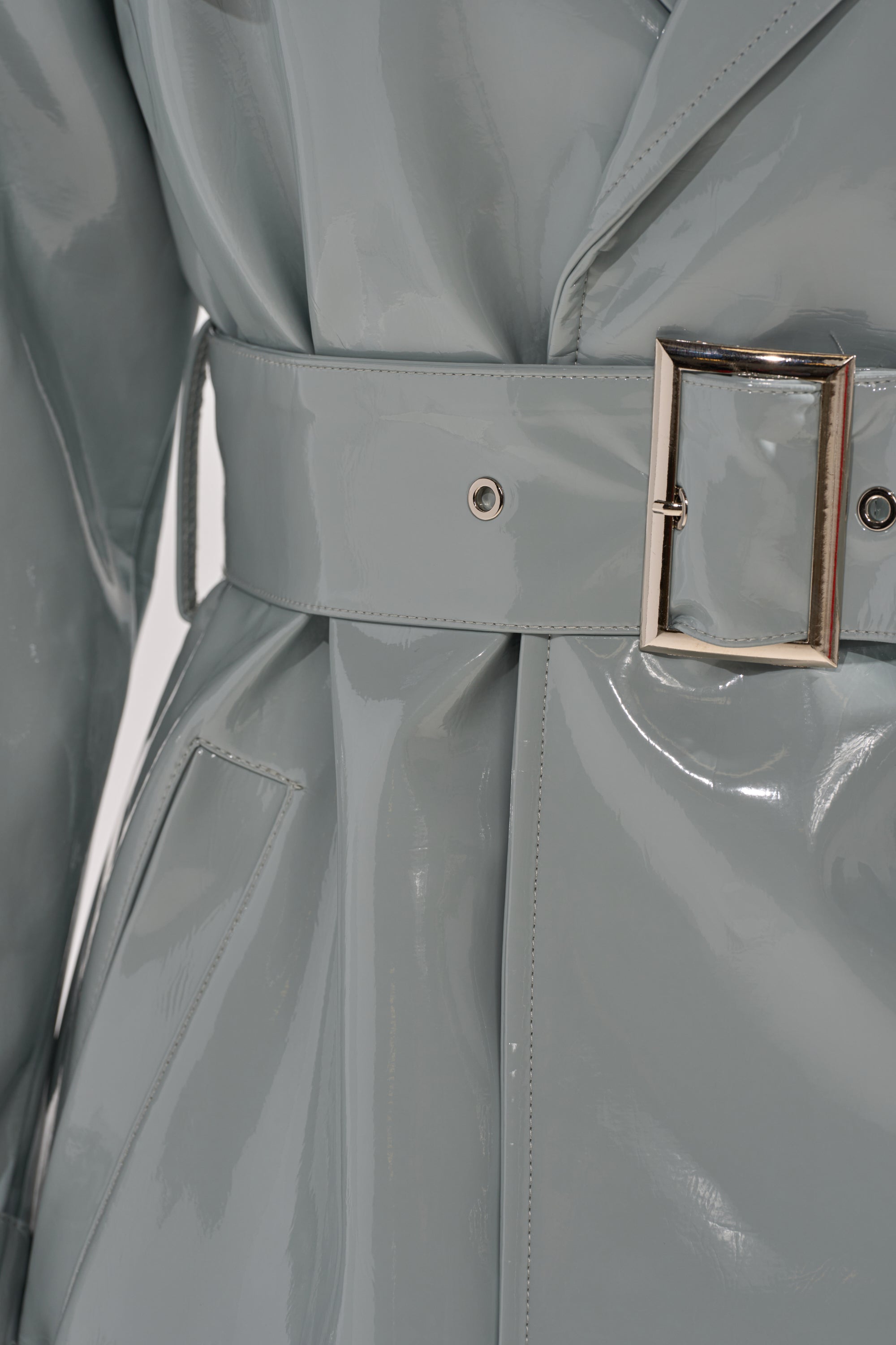 Close-up of the LOCKED AND LOADED PATENT TRENCH: a glossy light grey vegan patent leather coat with a matching belt and rectangular silver buckle, visible pocket stitching, and a chic, modern finish. 7