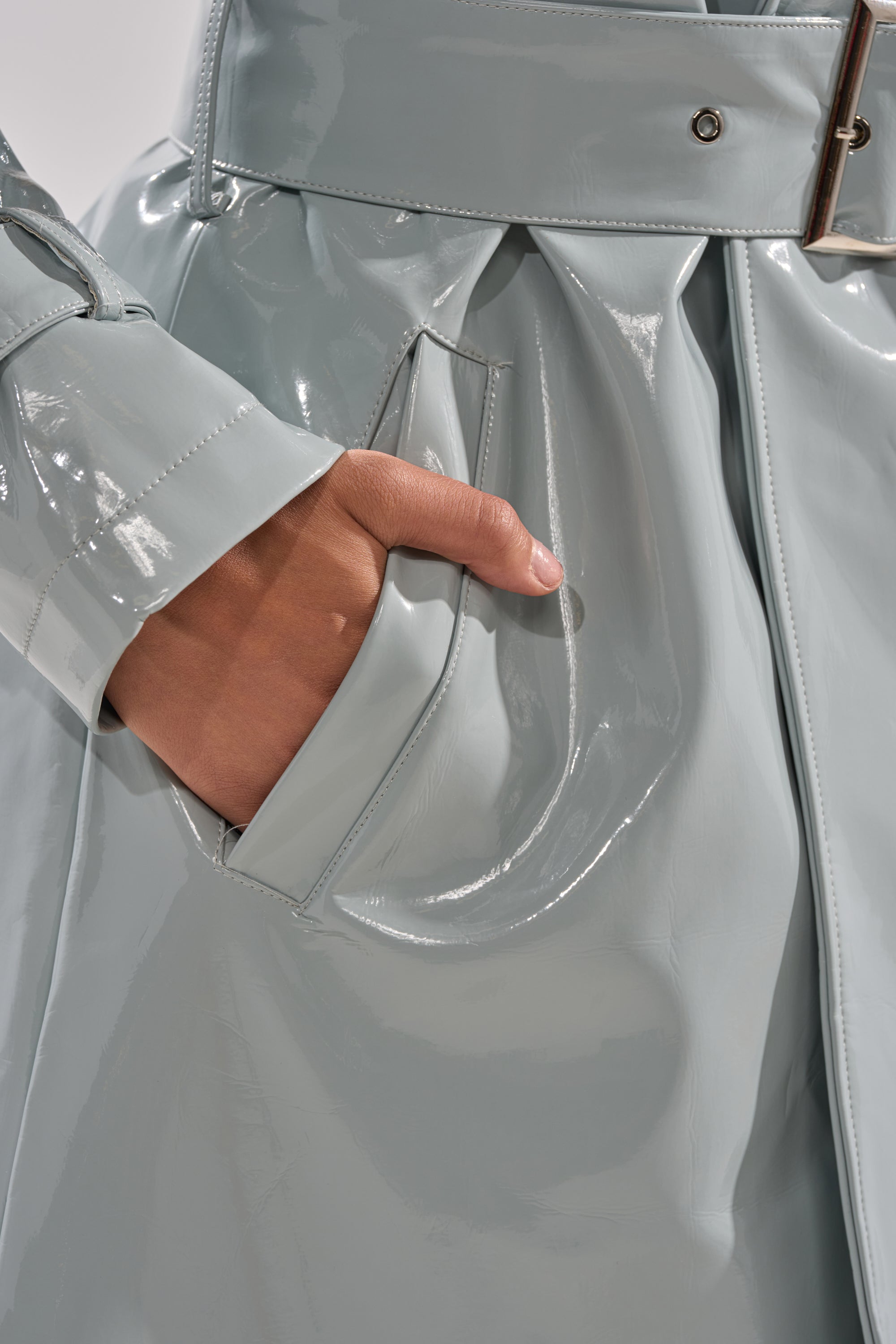 A close-up of a hand in the pocket of the LOCKED AND LOADED PATENT TRENCH, a shiny light gray vegan patent leather coat with a wide belt and glossy, reflective finish. 8