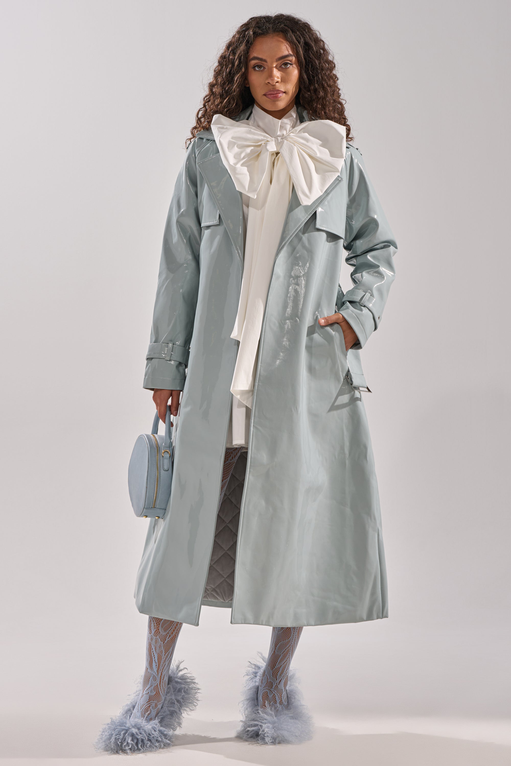 A woman wears the LOCKED AND LOADED PATENT TRENCH in shiny light blue vegan leather, with a large white neck bow, patterned blue tights, fluffy blue shoes, and a round blue handbag against a plain background. 1