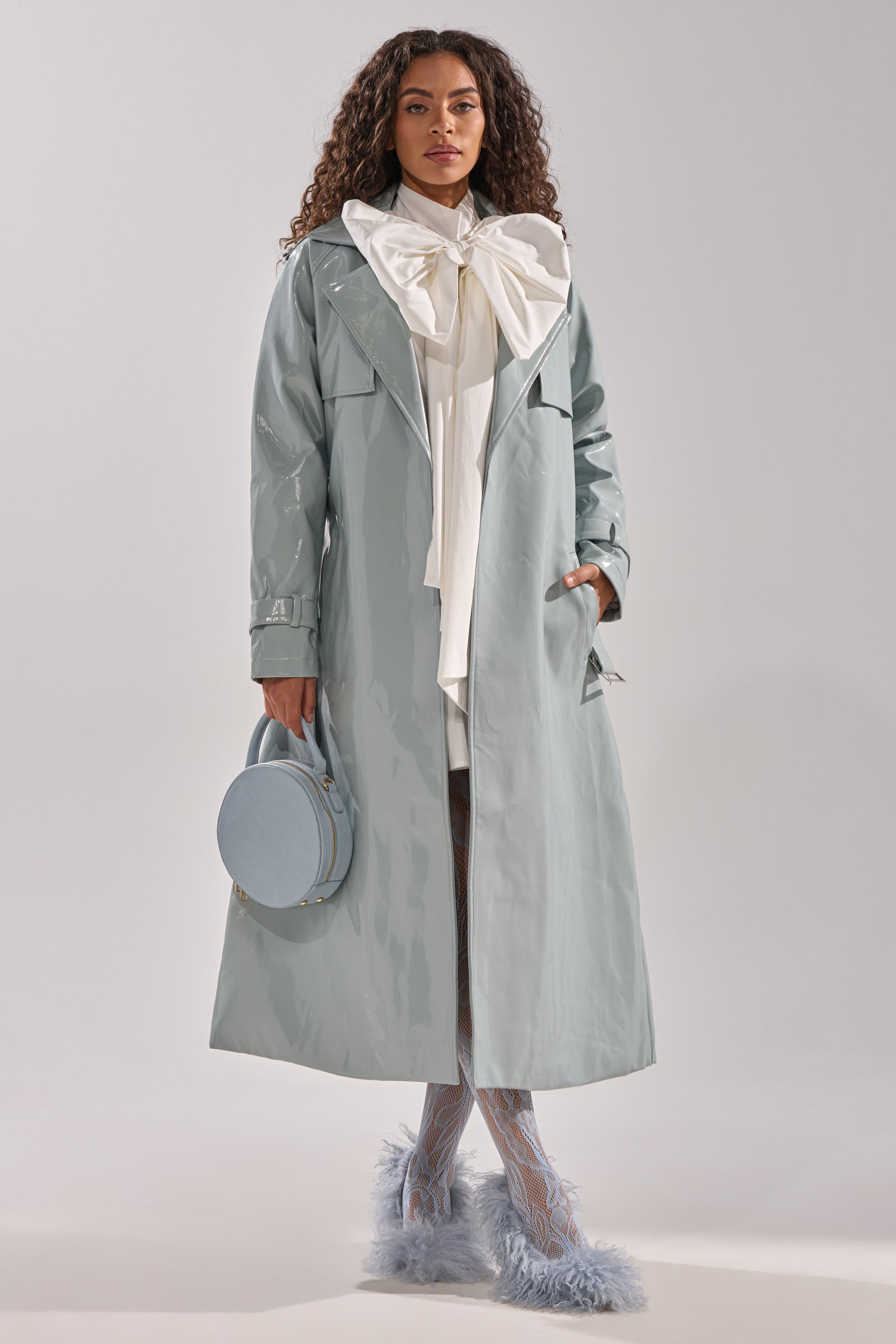 A woman with long curly hair and a confident look wears the shiny pale blue LOCKED AND LOADED PATENT TRENCH, a large white bow-tie blouse, patterned tights, fluffy blue shoes, and carries a round blue handbag. 2