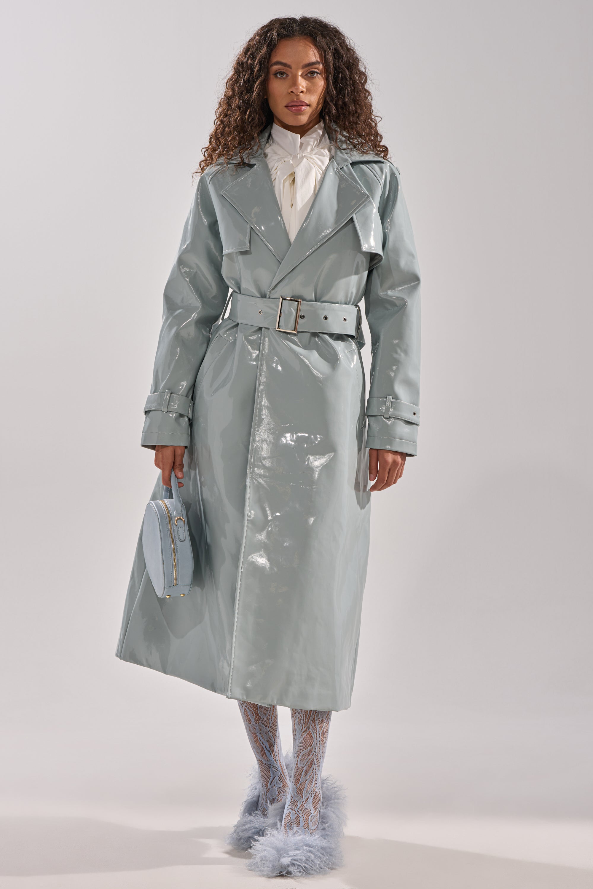 A woman wears the LOCKED AND LOADED PATENT TRENCH in shiny light blue with a belt, styled with a white blouse, patterned tights, fluffy blue shoes, and a matching handbag, standing against a plain background. 3
