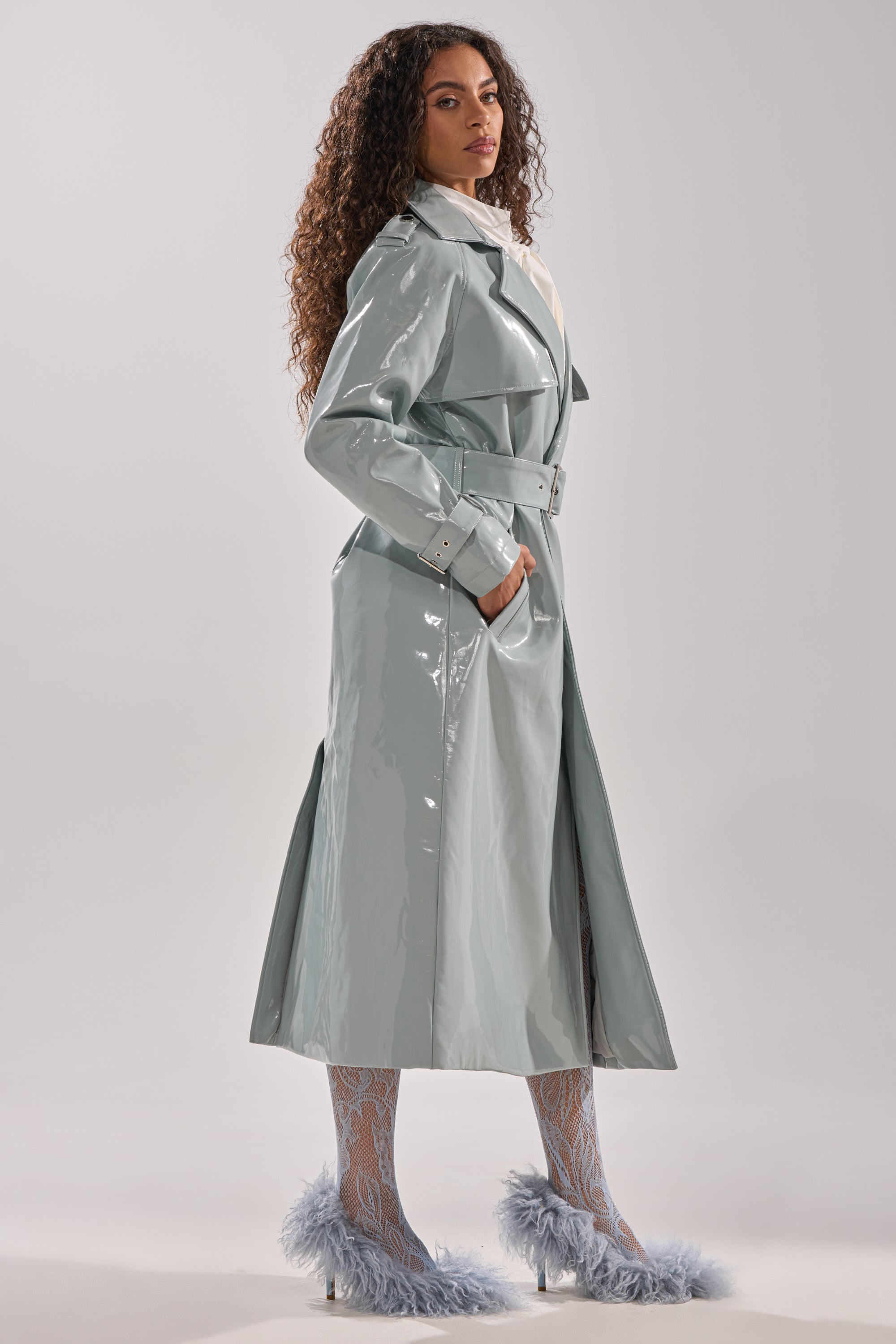 A woman with long curly hair stands sideways against a plain background, her left hand in her pocket, wearing the LOCKED AND LOADED PATENT TRENCH belted at the waist, paired with patterned tights and feather-trimmed heels. 4