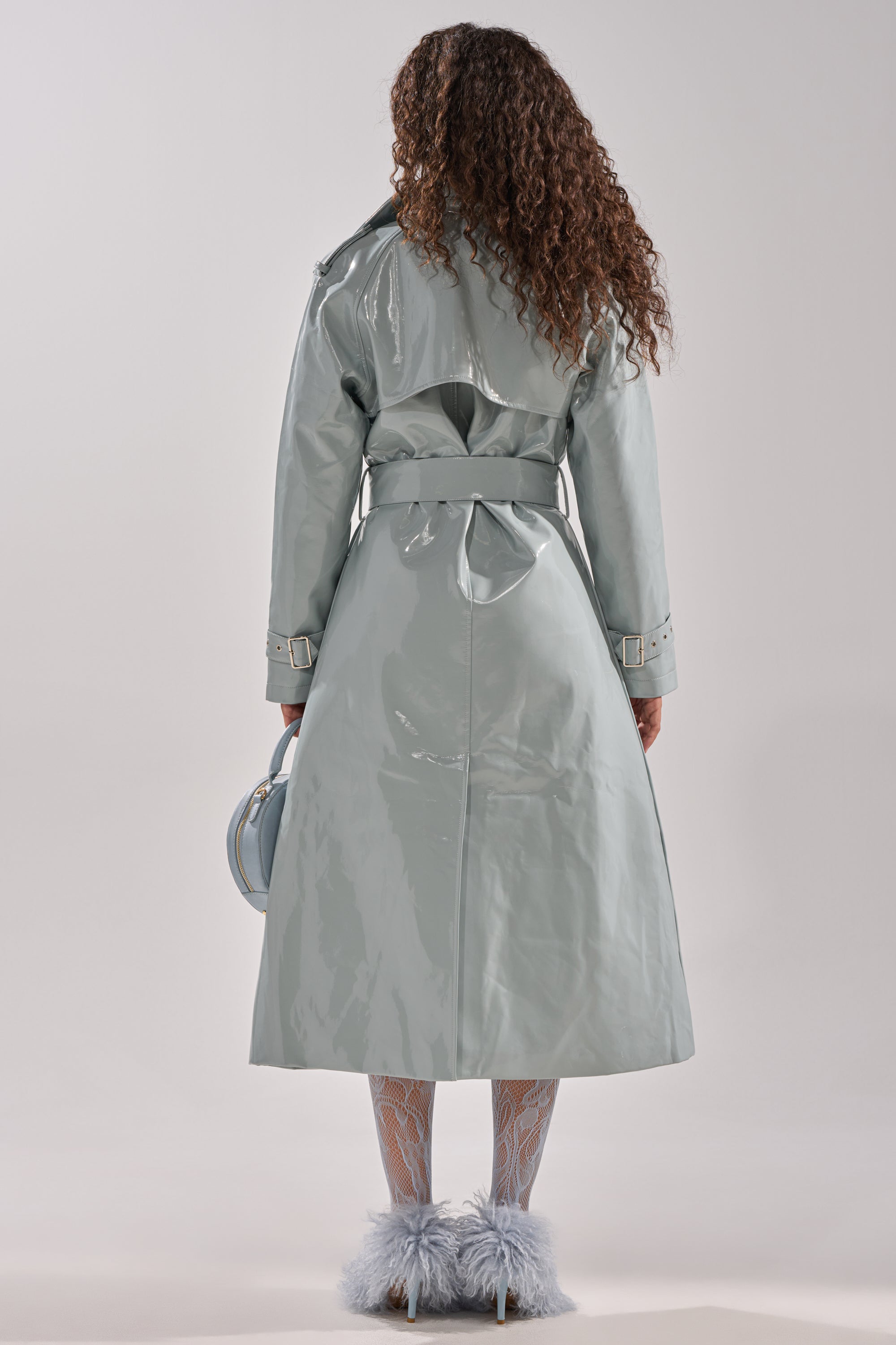 A person with long curly hair wears the LOCKED AND LOADED PATENT TRENCH in shiny light gray, patterned white tights, and fluffy gray shoes. Shown from the back, they hold a small round bag against a plain background. 5