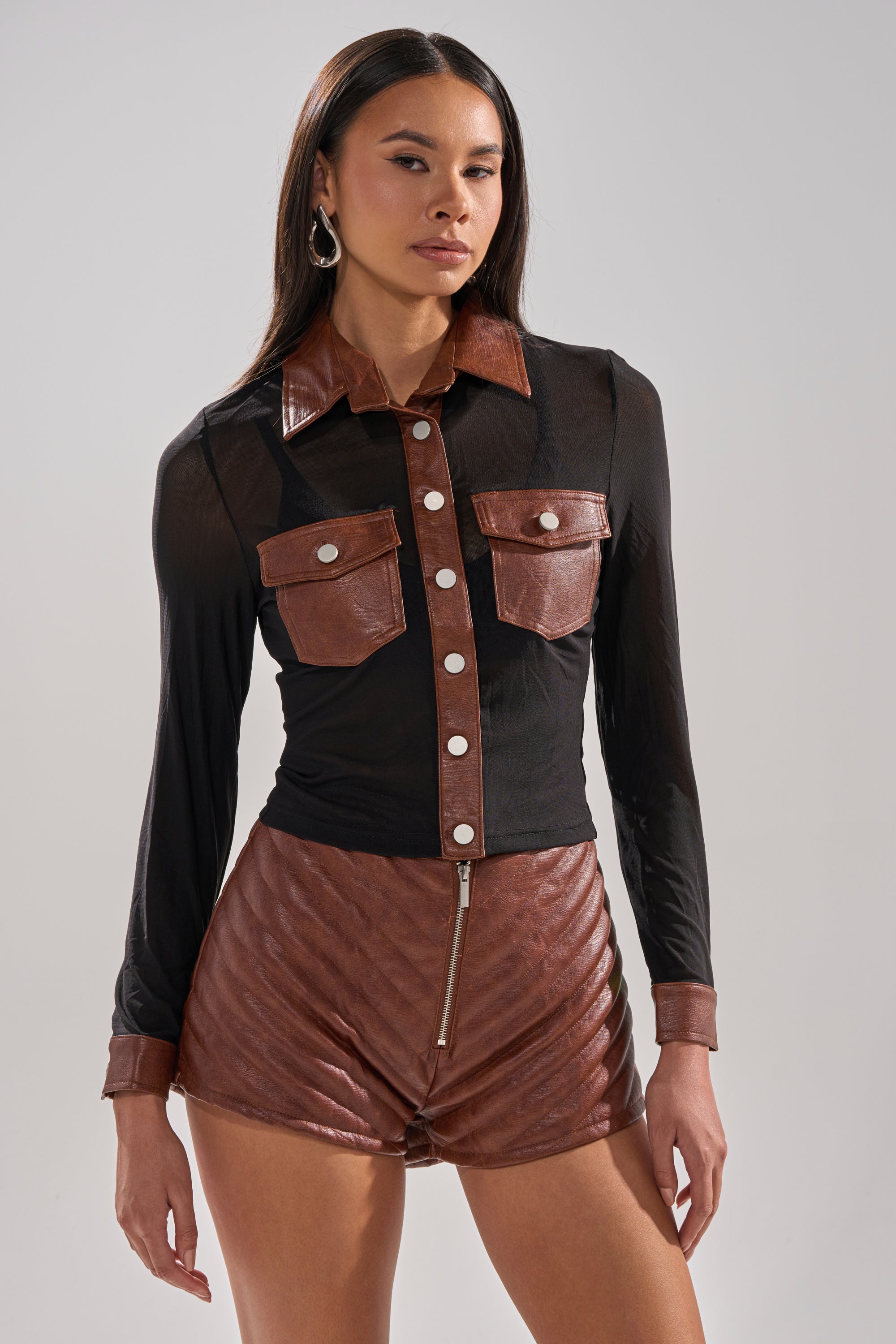A woman faces forward, wearing the GALAXY MESH BUTTON DOWN WITH FAUX LEATHER TRIM, featuring silver buttons, paired with matching brown shorts. She has long, straight hair and hoop earrings.