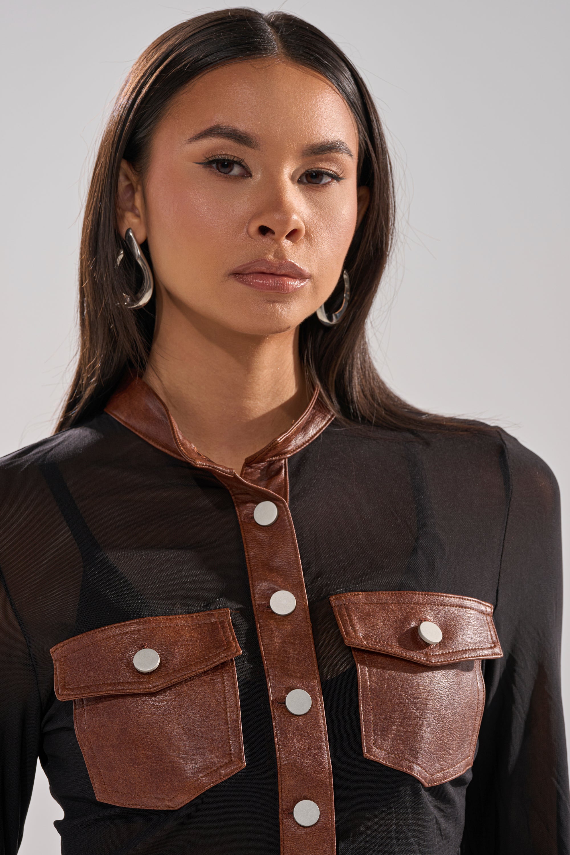 A woman with long dark hair wears silver hoop earrings and the GALAXY MESH BUTTON DOWN WITH FAUX LEATHER TRIM, a sheer top with black and brown trim, pockets, and silver buttons, set against a neutral background.
