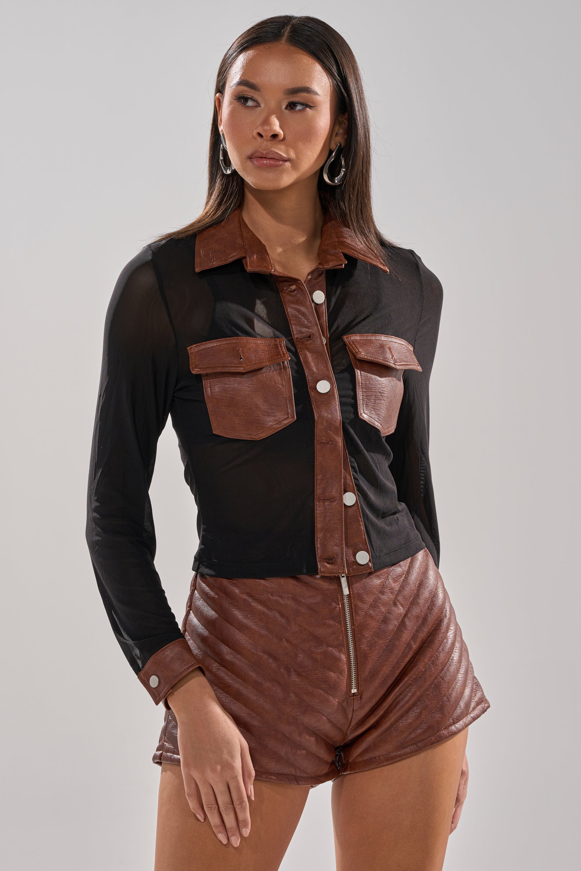 A woman wears the GALAXY MESH BUTTON DOWN WITH FAUX LEATHER TRIM and brown quilted leather shorts, standing against a plain light background.