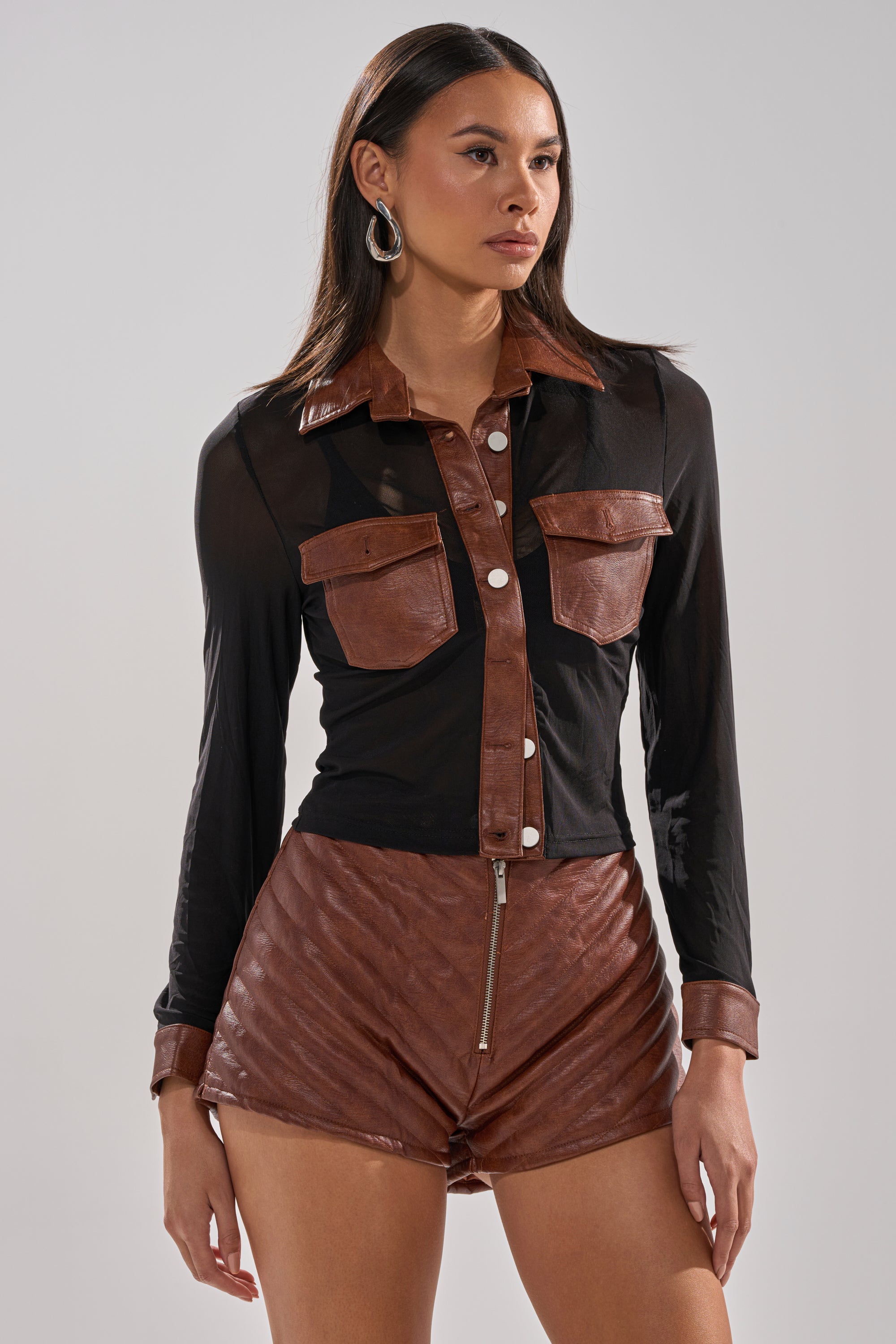 A woman wears the GALAXY MESH BUTTON DOWN WITH FAUX LEATHER TRIM, featuring black sheer mesh and faux leather on collar, cuffs, buttons, and chest pockets, paired with brown leather shorts, long straight hair, and large hoop earrings.