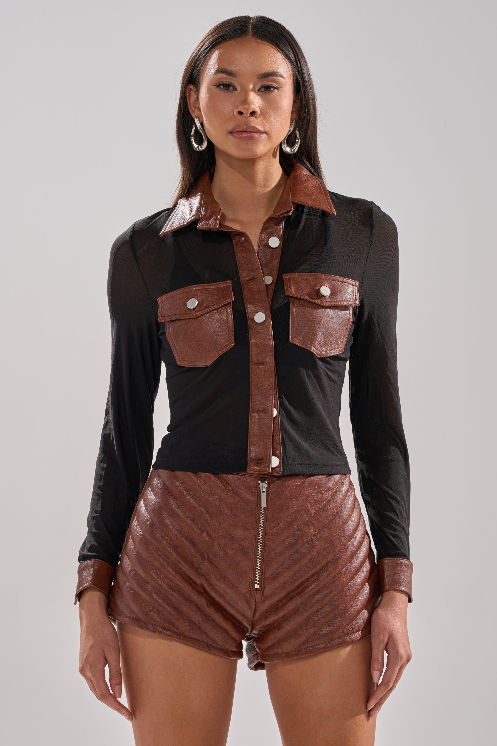 A woman faces forward wearing the GALAXY MESH BUTTON DOWN WITH FAUX LEATHER TRIM, paired with high-waisted, quilted brown leather shorts with a front zipper.
