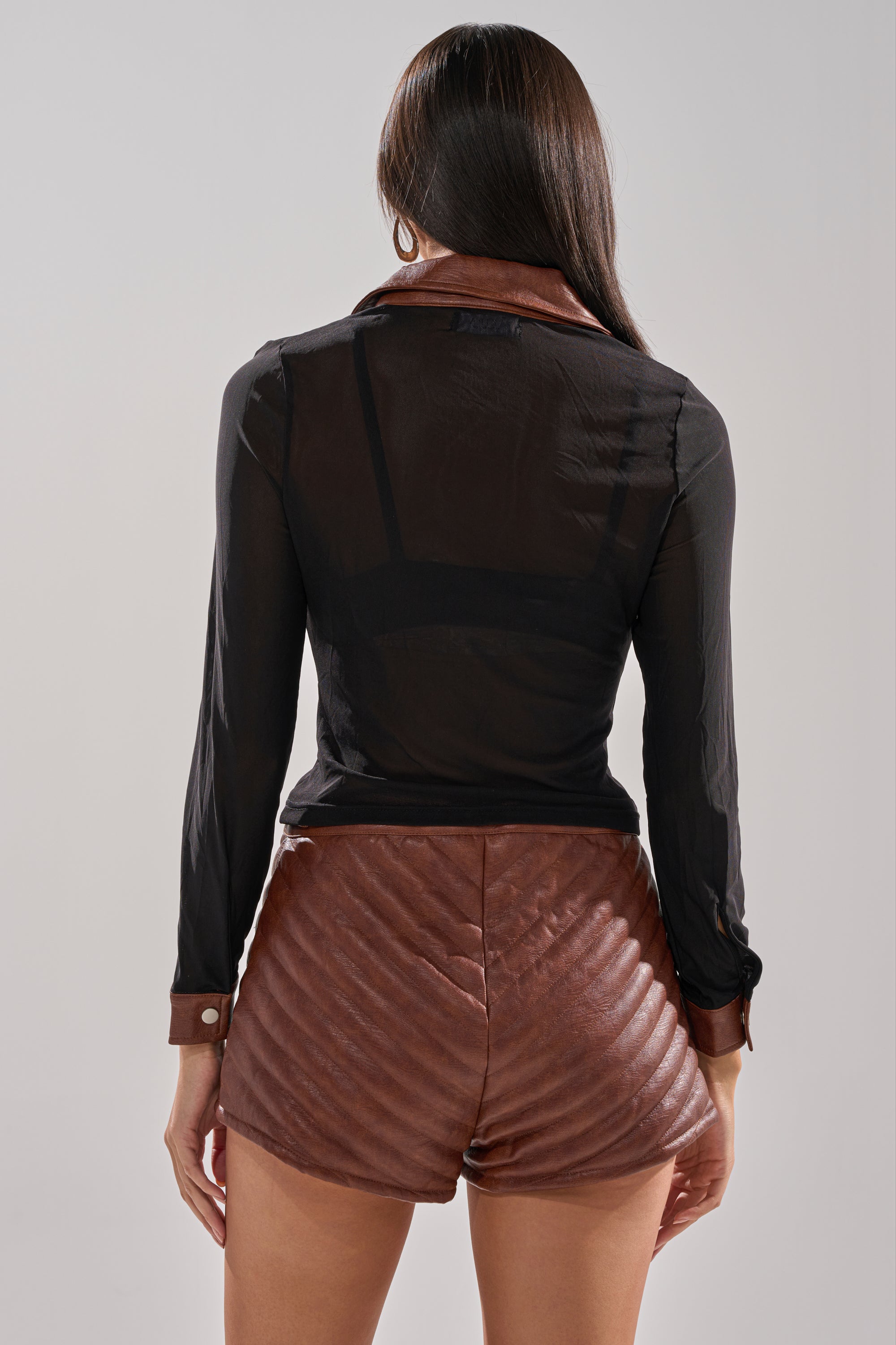 A person with long dark hair is seen from the back, wearing the GALAXY MESH BUTTON DOWN WITH FAUX LEATHER TRIM and high-waisted brown quilted shorts against a plain, light-colored background.