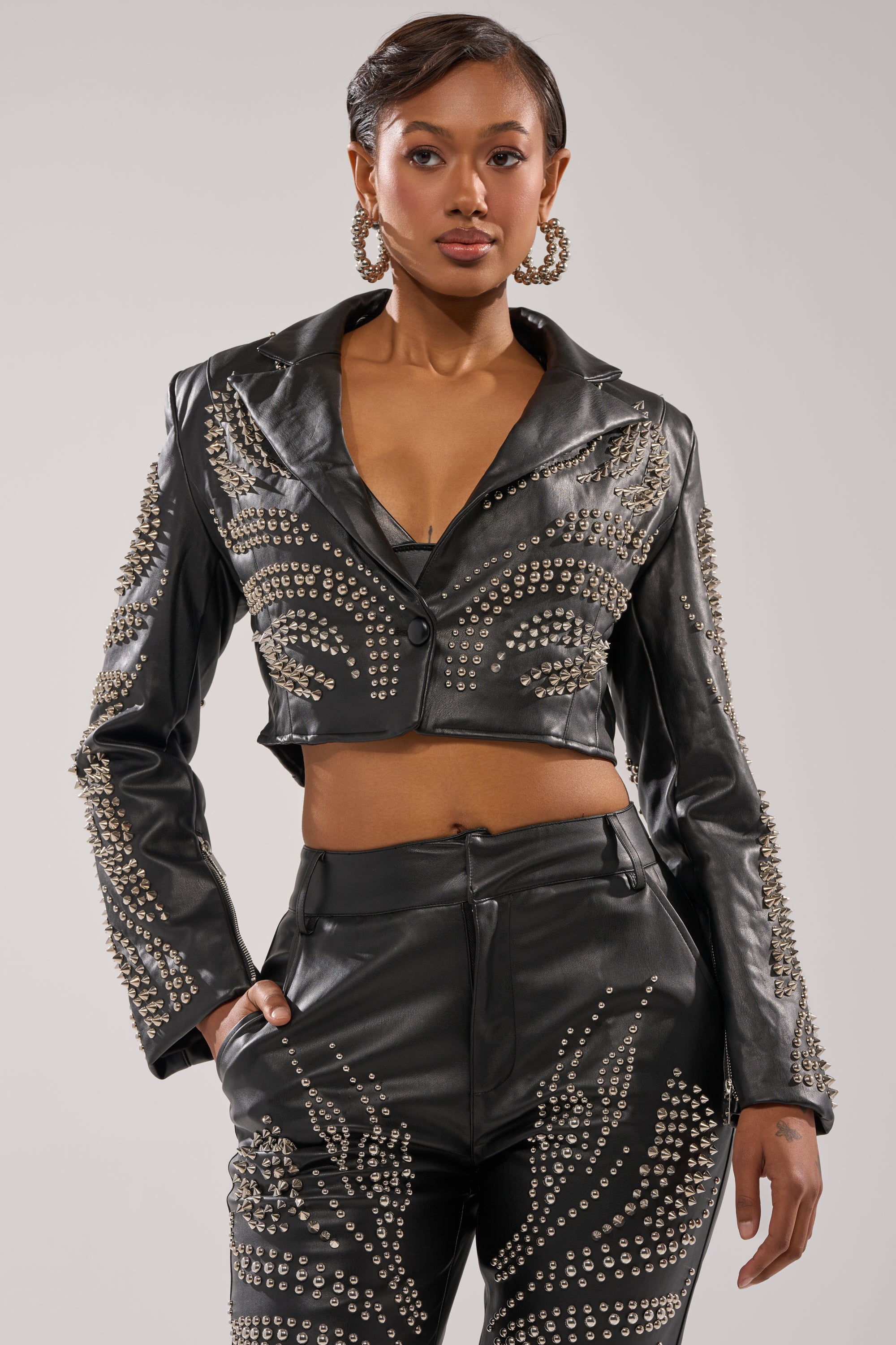 A woman poses confidently in the GONE BAD STUDDED BLAZER, a cropped faux leather piece with intricate silver stud patterns, paired with matching pants. She accessorizes with large hoop earrings and has her hair neatly styled back. 1