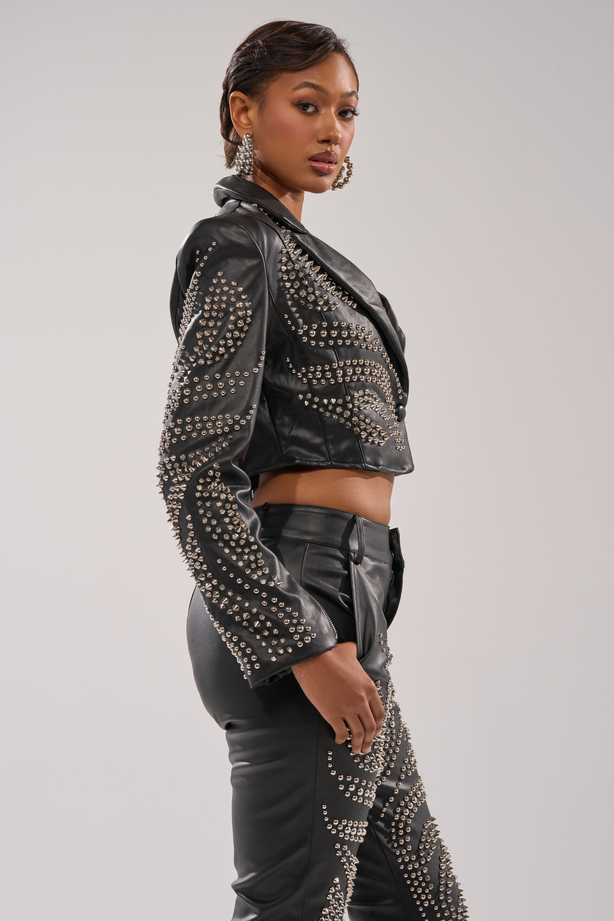 A woman models the GONE BAD STUDDED BLAZER—a black, fitted faux leather blazer with silver studs—paired with matching pants. She wears hoop earrings and has her hair styled back, set against a plain light background. 3