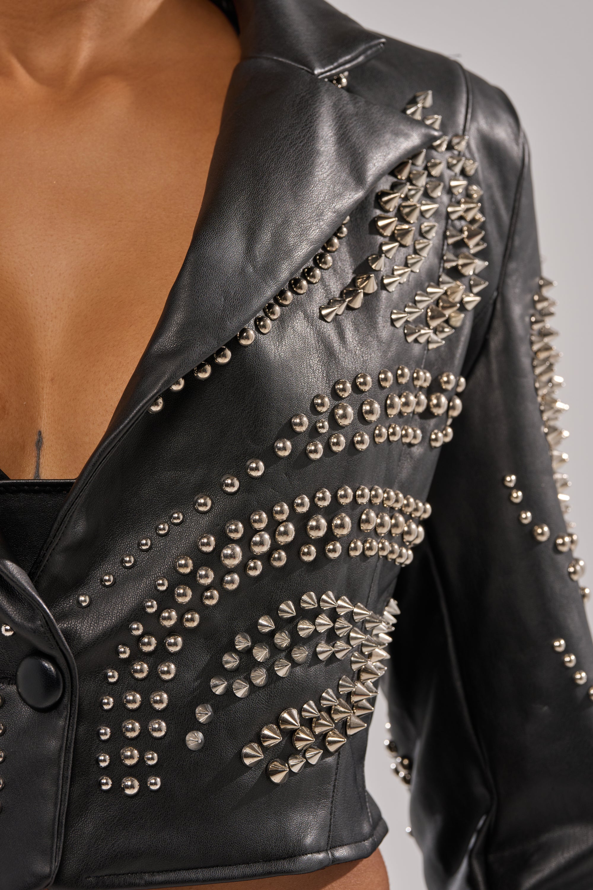 A close-up of a person wearing the GONE BAD STUDDED BLAZER, featuring silver studs and spikes in swirling patterns. The blazer is slightly open at the chest, revealing skin and a black garment underneath. 7