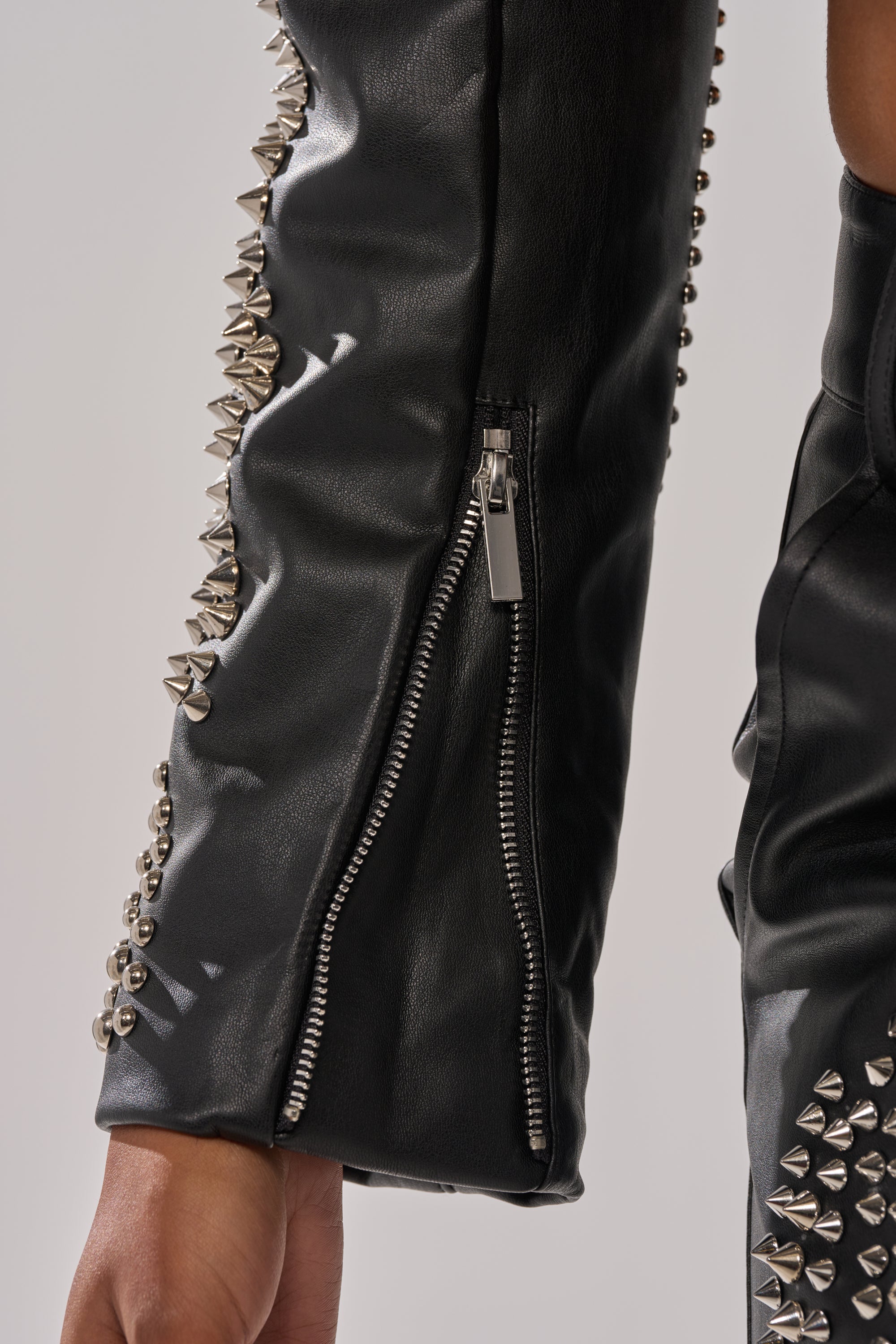 A close-up of the GONE BAD STUDDED BLAZER sleeve in black faux leather, featuring silver spikes and a visible zipper, with a person's hand partially visible at the bottom. 9