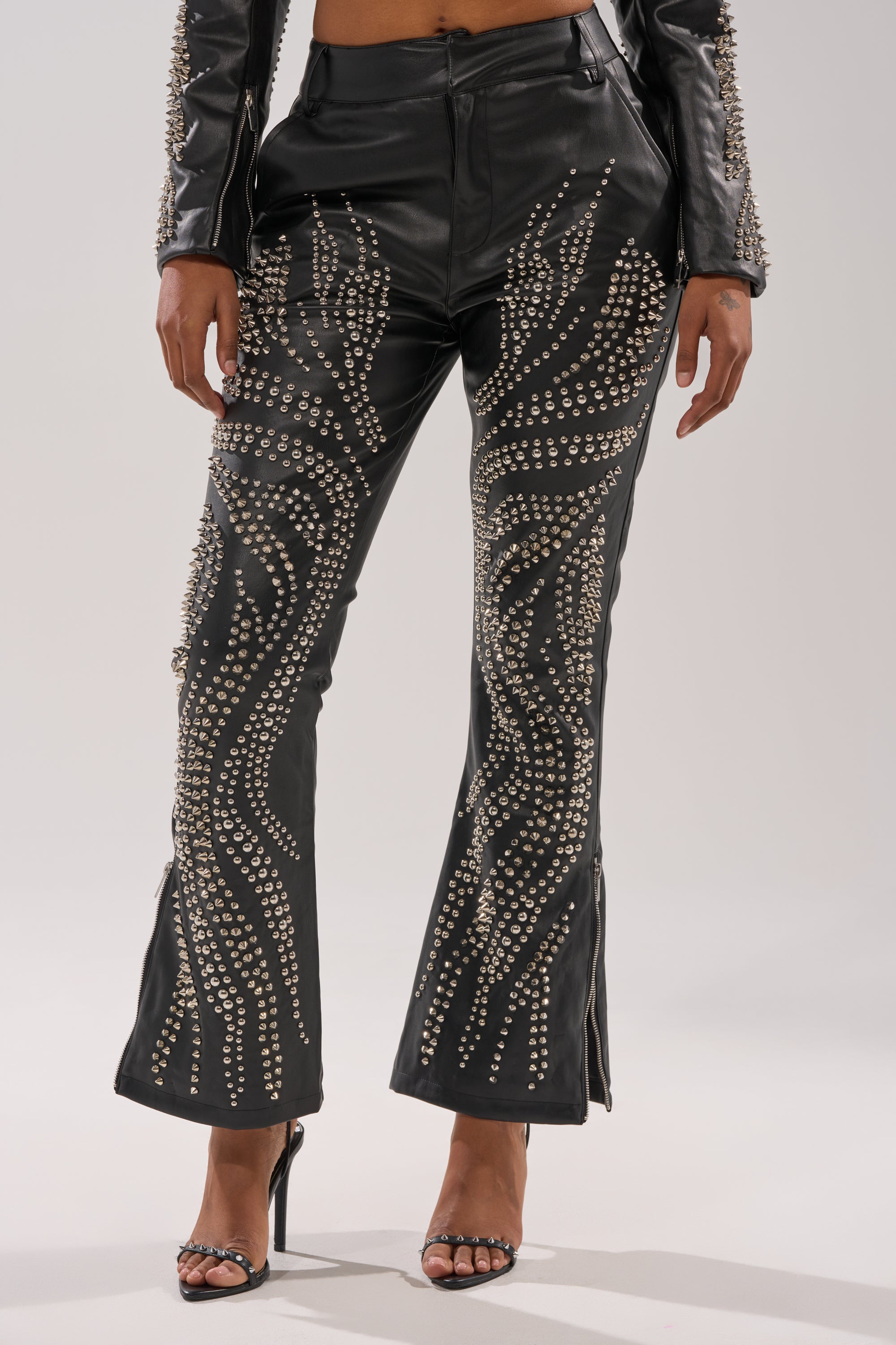 A person wears GONE BAD STUDDED PANT featuring swirling silver studs, paired with a matching studded jacket and open-toed black heels. The background is plain and light-colored.