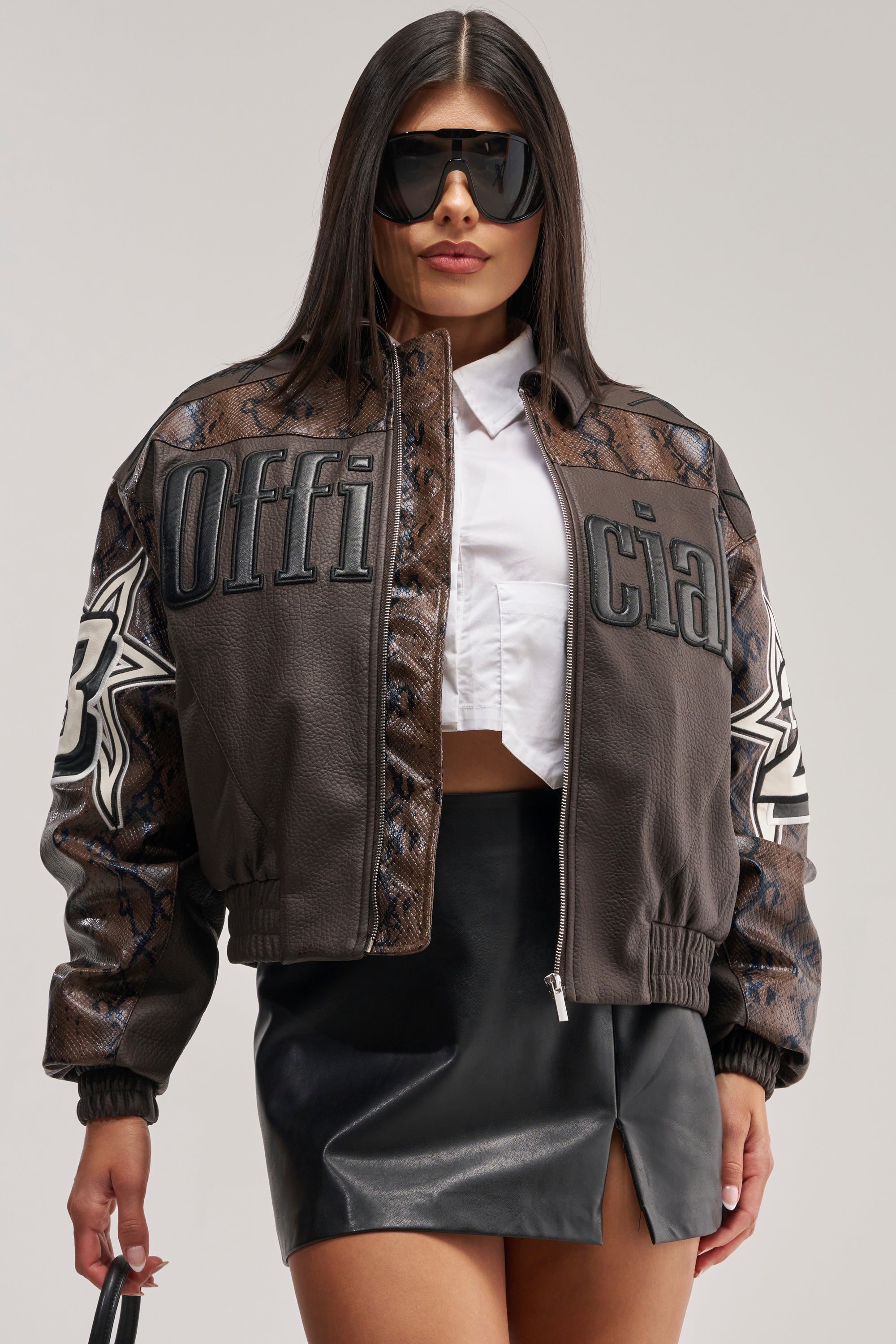 A woman stands confidently against a plain background wearing the MAKE IT OFFICIAL VARSITY BOMBER—a cropped leather jacket with "Official" text and sleek snakeskin paneling—paired with a white cropped shirt, a black leather mini skirt, and large sunglasses.