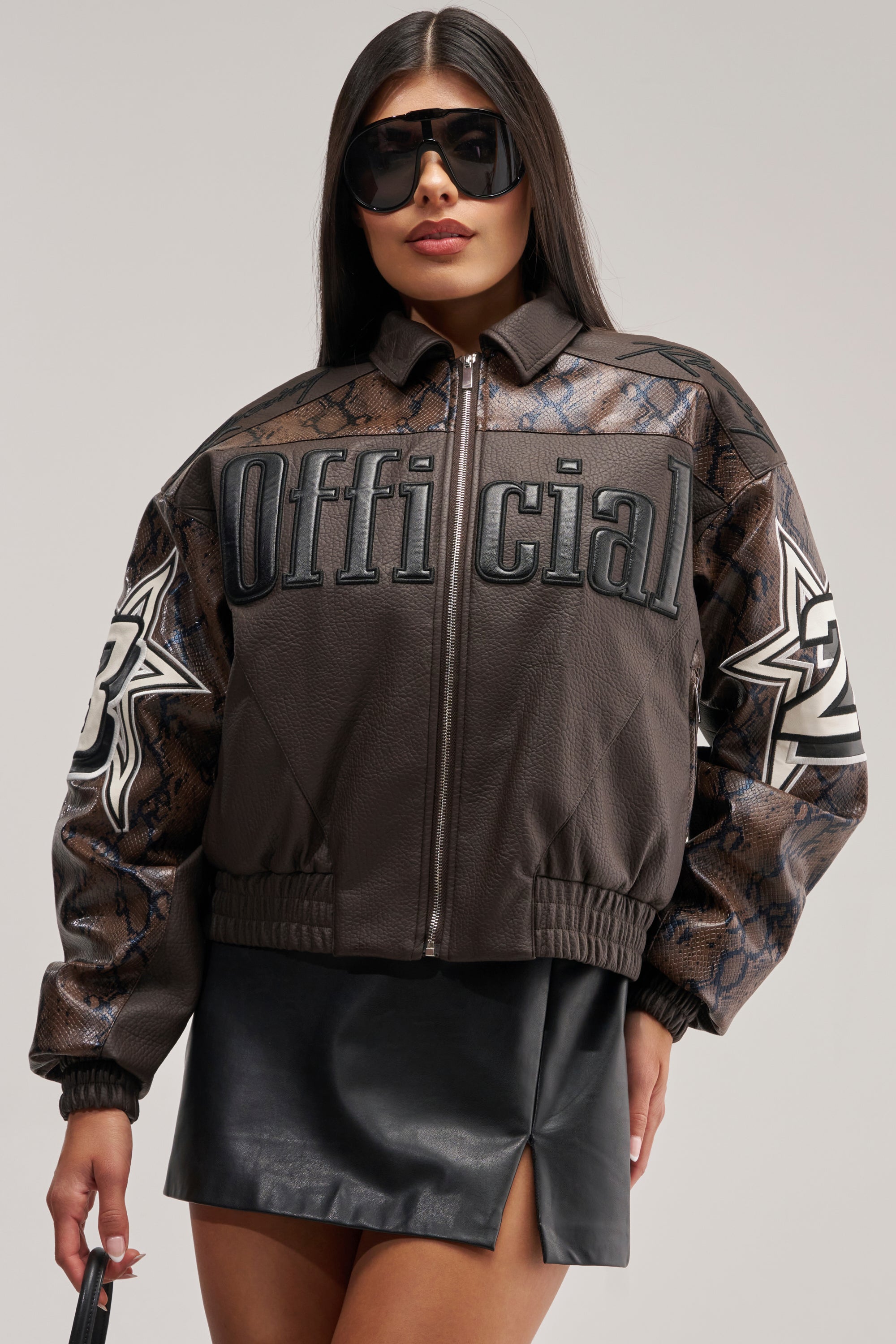 A woman models the MAKE IT OFFICIAL VARSITY BOMBER—an oversized dark jacket featuring bold snakeskin-patterned sleeves, star patches, and "Official" text—styled with a black mini skirt and statement sunglasses, posing confidently against a simple backdrop.
