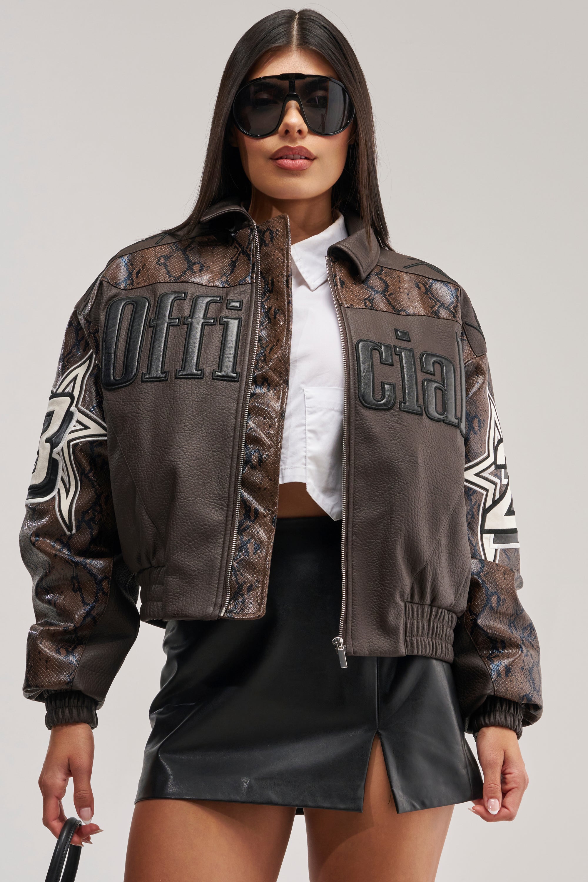 A woman wearing black sunglasses, the MAKE IT OFFICIAL VARSITY BOMBER featuring dark brown faux leather with snakeskin paneling and "Official" text alongside white star designs, a white crop top, and a black leather mini skirt poses confidently against a plain background.