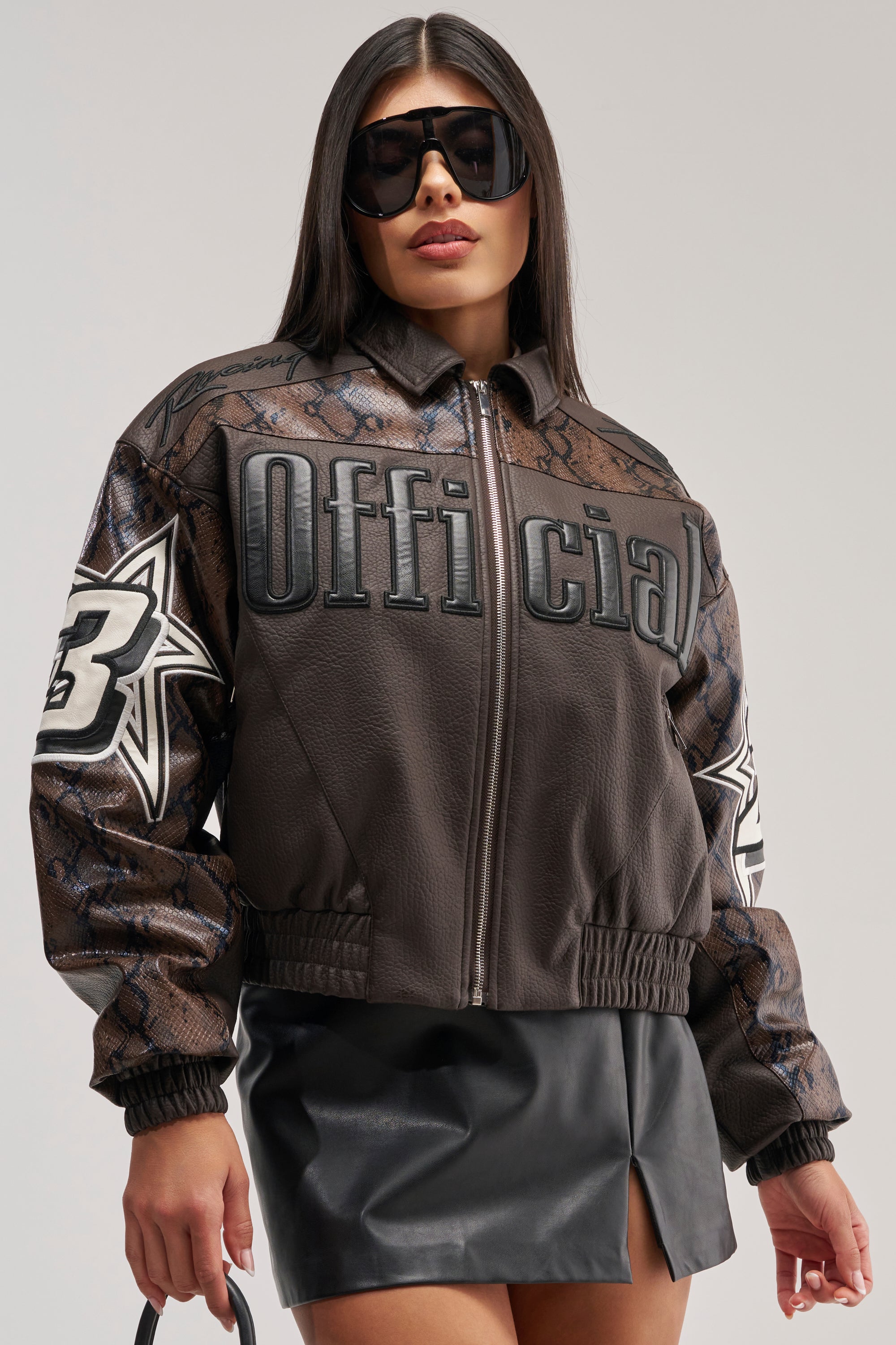 A woman wearing oversized sunglasses and the MAKE IT OFFICIAL VARSITY BOMBER—a brown faux leather jacket featuring "Official" text and star patches—paired with a black leather mini skirt, stands against a plain background.