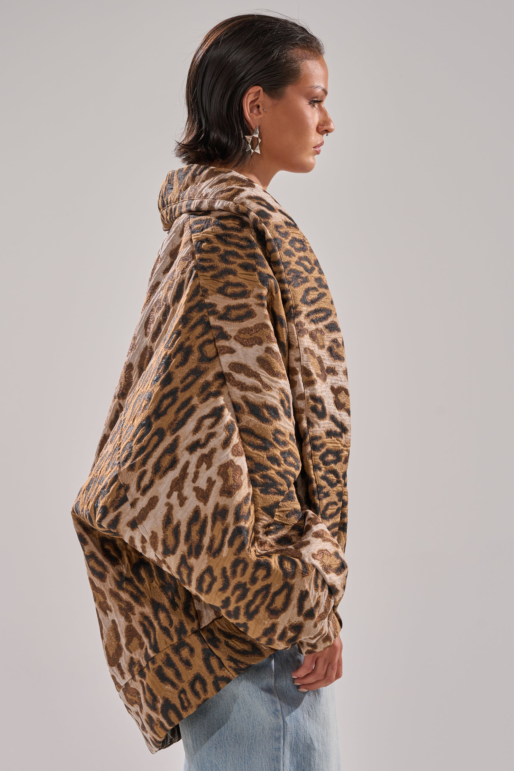 A person in profile wears the LOLA COCOON COAT IN JACQUARD—an oversized, hooded leopard print coat with a cocoon silhouette—paired with light blue jeans. A plain, neutral background accentuates the coat’s bold pattern and unique shape.