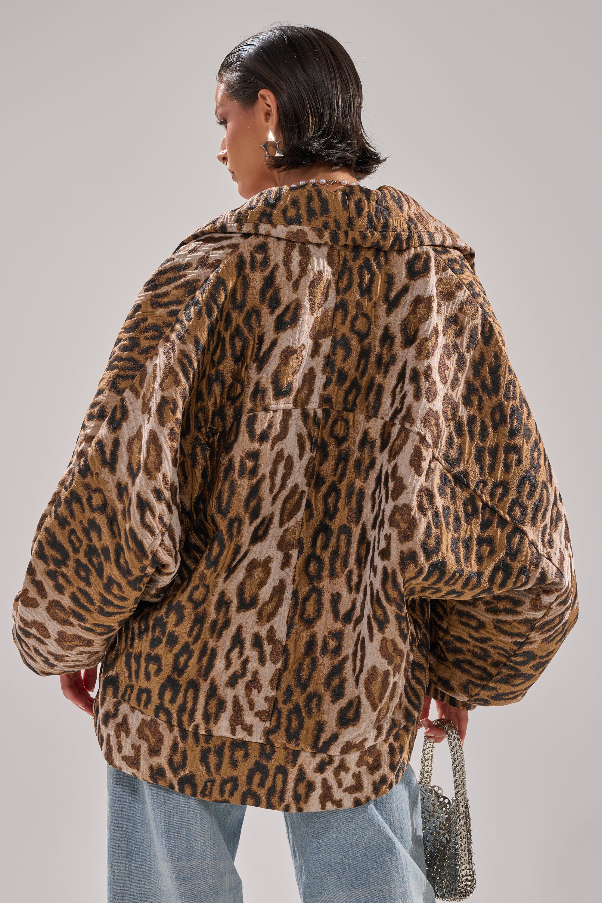 A person with short dark hair wears the LOLA COCOON COAT IN JACQUARD, a loose, oversized leopard print piece, paired with light blue jeans and holding a small metallic handbag. The bold animal print of the coat is showcased as they face away.