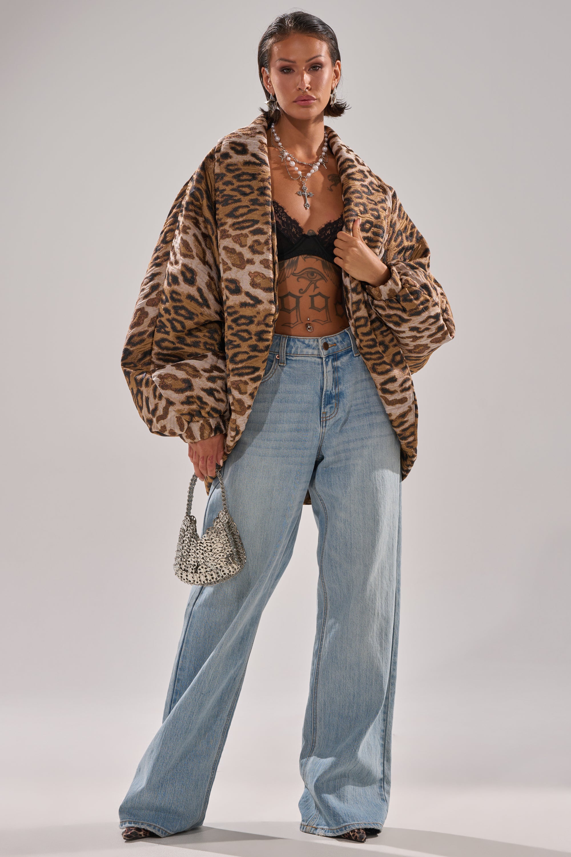 A woman in the LOLA COCOON COAT IN JACQUARD, black lace bralette, wide-leg light blue jeans, and leopard print shoes carries a beaded handbag. She has short hair, visible tattoos, and layered jewelry.
