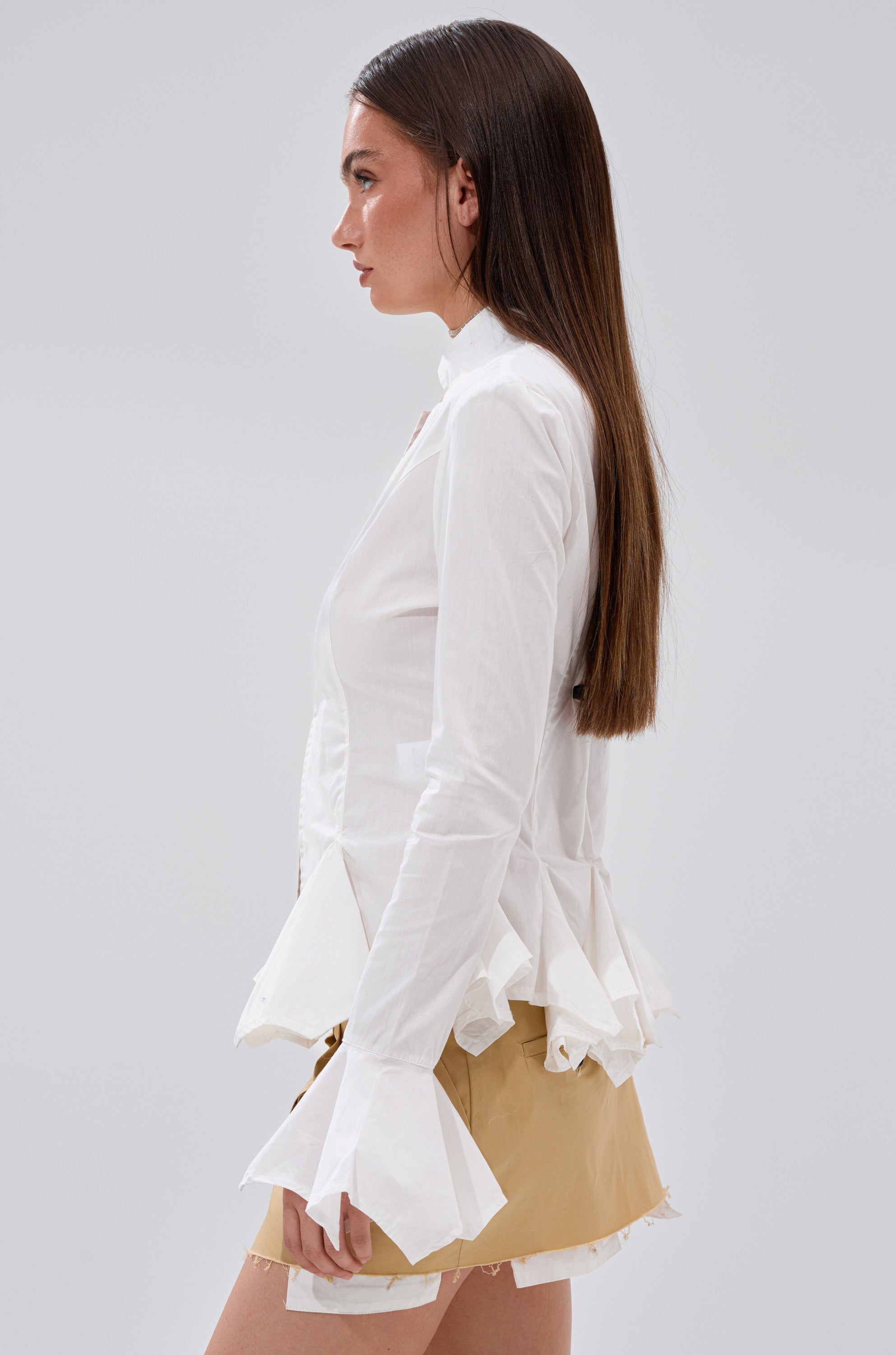A woman with long straight brown hair is shown in profile, wearing the ITS GOING TO BE ALRIGHT BUTTON DOWN blouse with flared cuffs and a ruffled hem, paired with a beige mini skirt against a plain light gray background.