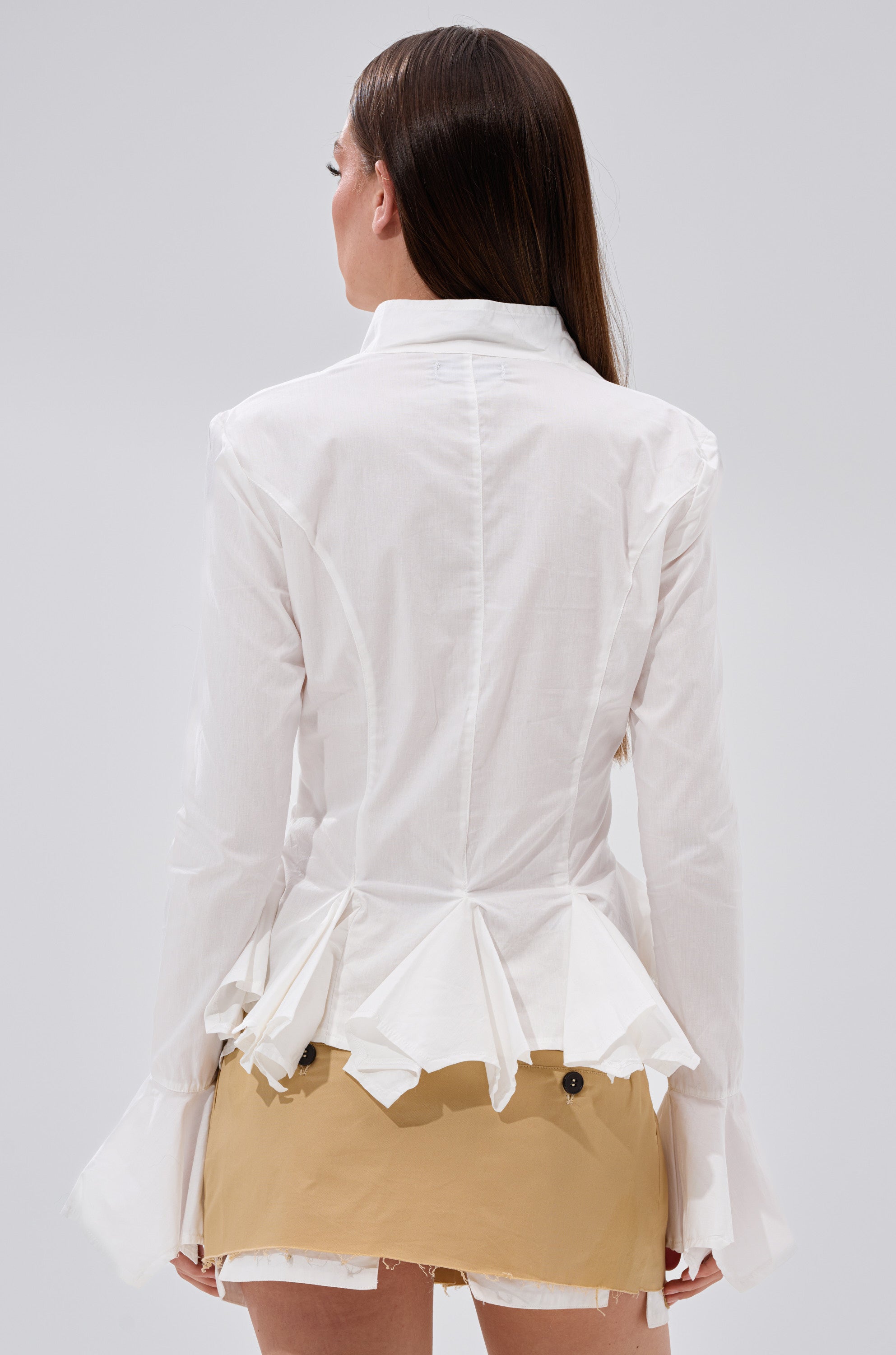 A woman with long brown hair is seen from behind, wearing the ITS GOING TO BE ALRIGHT BUTTON DOWN, a white peplum top with ruffled details and flared cuffs, paired with a tan skirt with prominent buttons and a light gray backdrop.