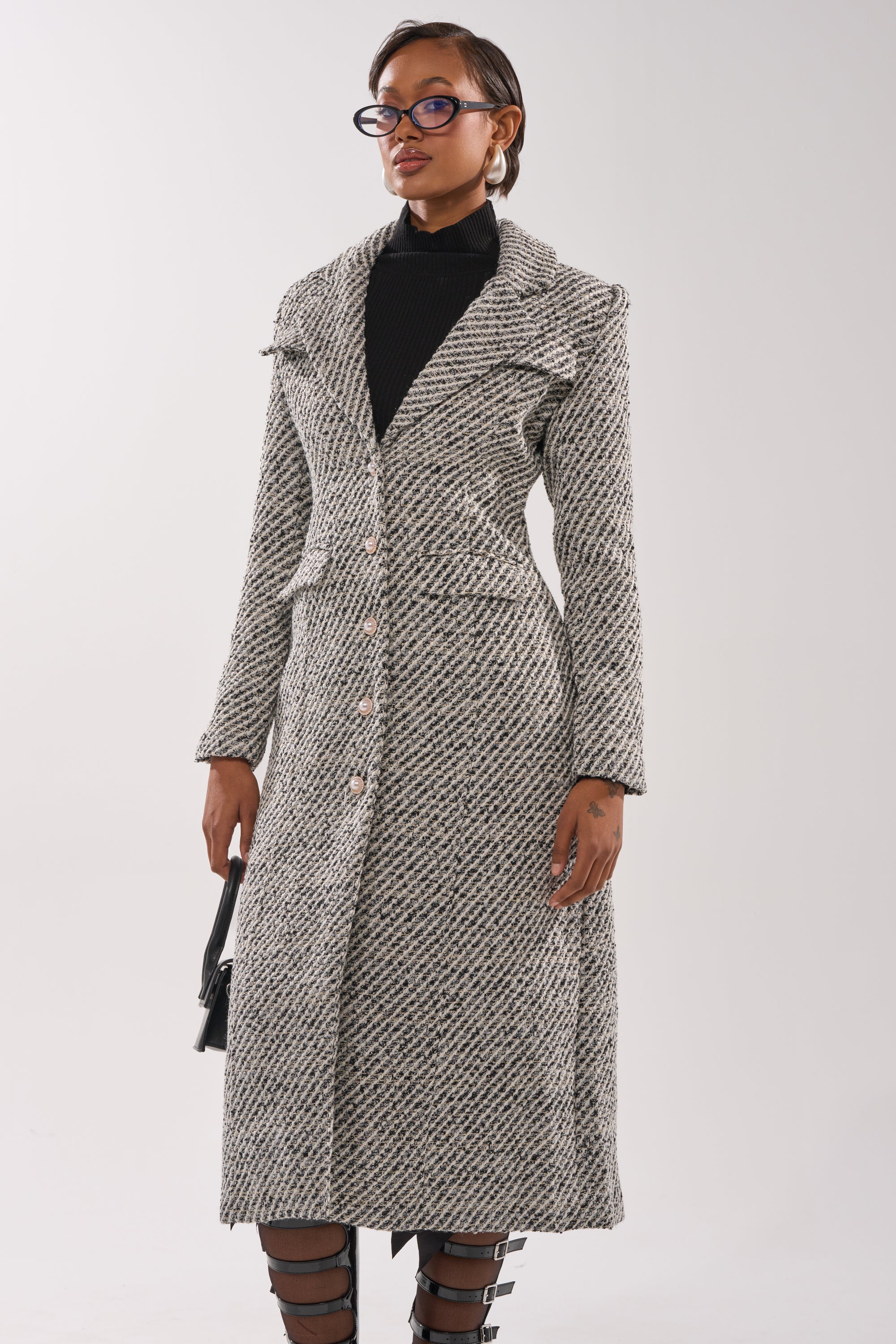 A woman with short hair and glasses stands against a plain background, wearing the SEVILLE TWEED TRENCH over a black turtleneck, paired with strappy black shoes and holding a small black handbag.