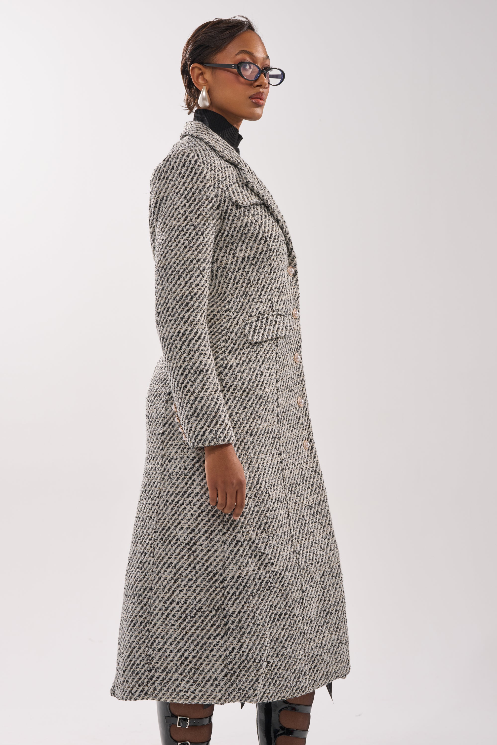 A woman in glasses and earrings models the SEVILLE TWEED TRENCH—a long, textured gray winter coat with a subtle pattern—paired with black boots, standing in profile against a plain light background.
