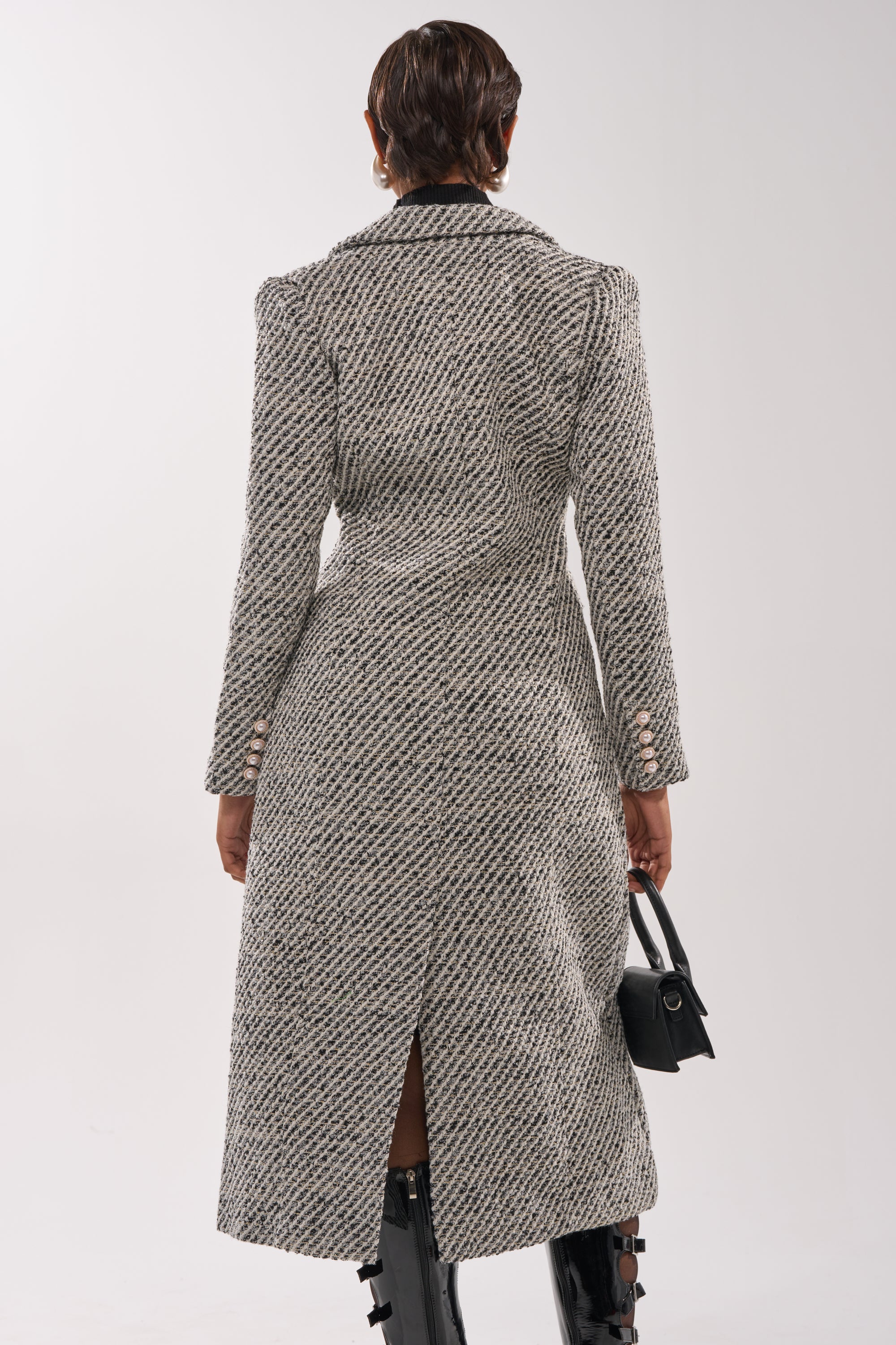 A person with short hair faces away, wearing the fitted gray SEVILLE TWEED TRENCH with buttoned sleeves, tall black boots, and holding a small black handbag against a plain light background.