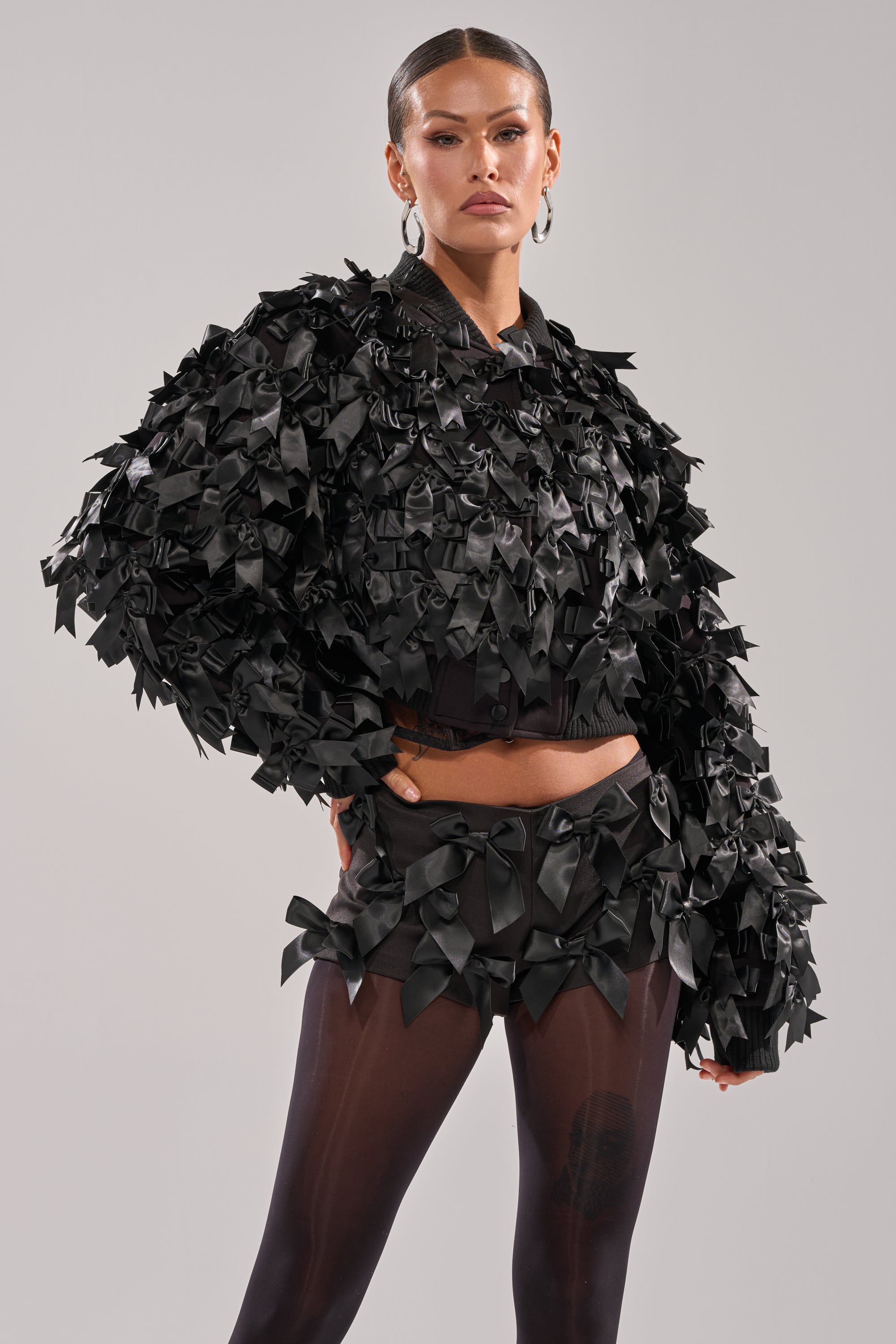A woman poses confidently in the black BOW DOWN SATIN BOMBER by AKIRA Label, featuring shiny textured bows. She stands against a light background, wearing sheer black tights, matching earrings, and her hair pulled back sleekly. 0