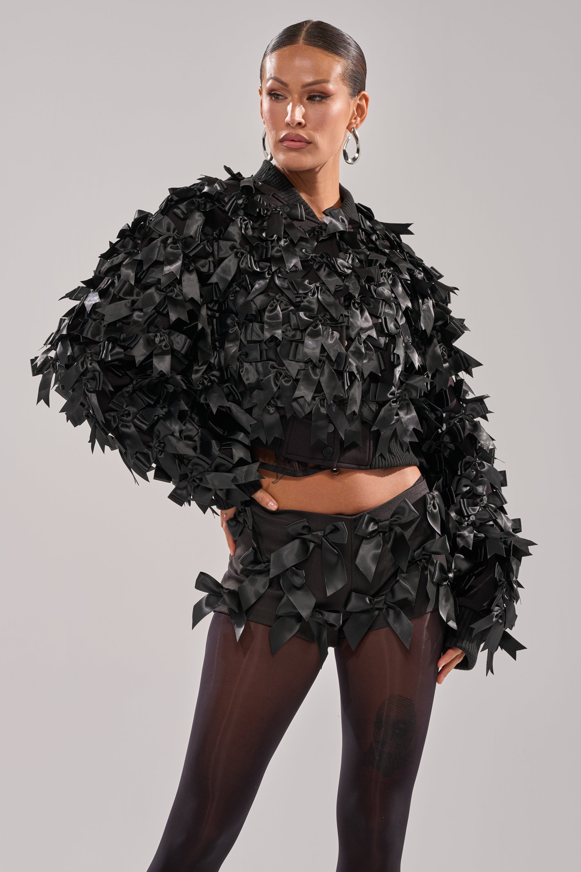 A woman poses confidently in the AKIRA Label BOW DOWN SATIN BOMBER—an all-black outfit with small satin bows, voluminous cropped bomber top, matching shorts, and sheer black tights against a plain light background. 3