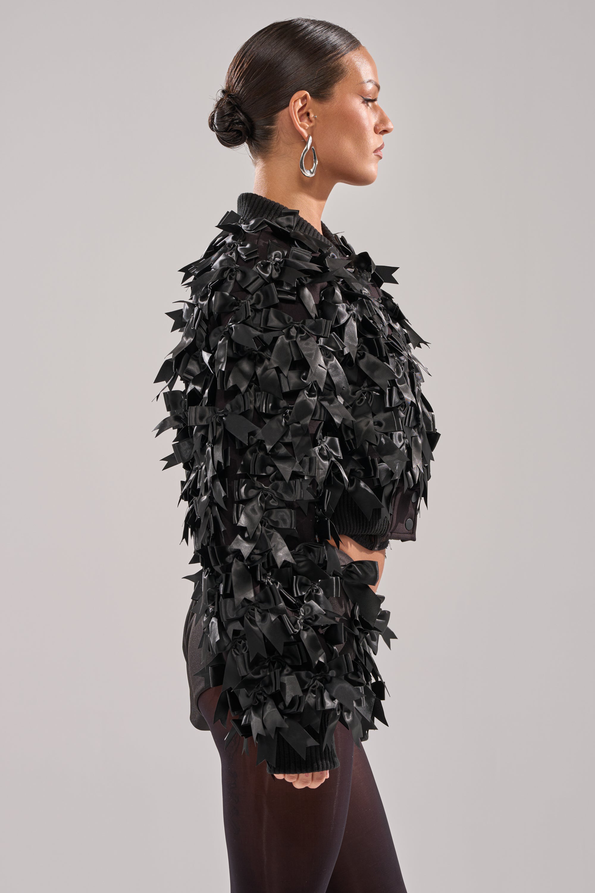 A woman with slicked-back hair wears large hoop earrings and the BOW DOWN SATIN BOMBER, featuring numerous ribbon bows, while standing in profile against a neutral background. 4