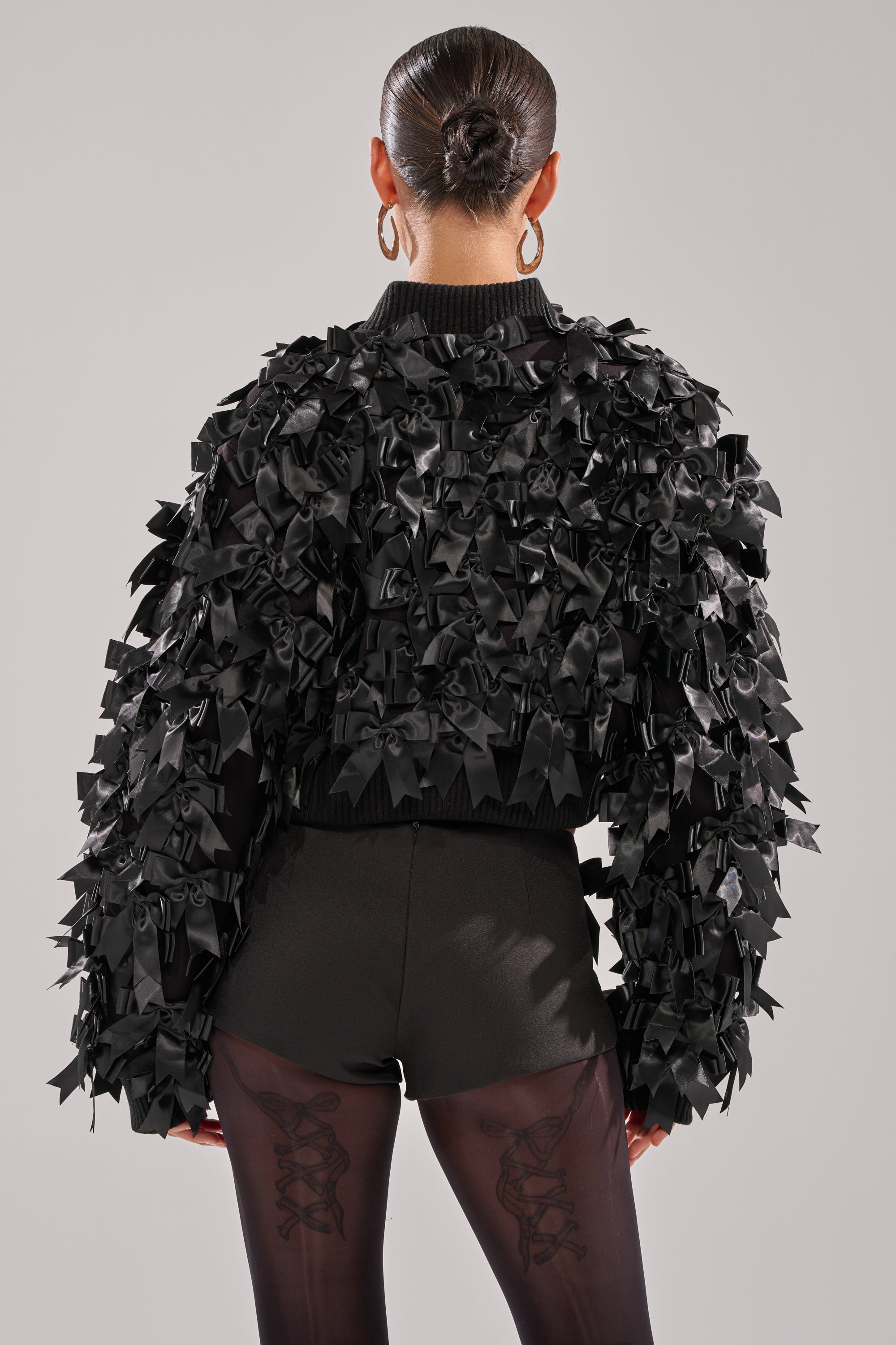 Wearing the BOW DOWN SATIN BOMBER with black bow appliques, a person with slicked-back hair and hoop earrings pairs it with sheer black tights decorated with ribbon patterns on the back, posing with their back to the camera. 5