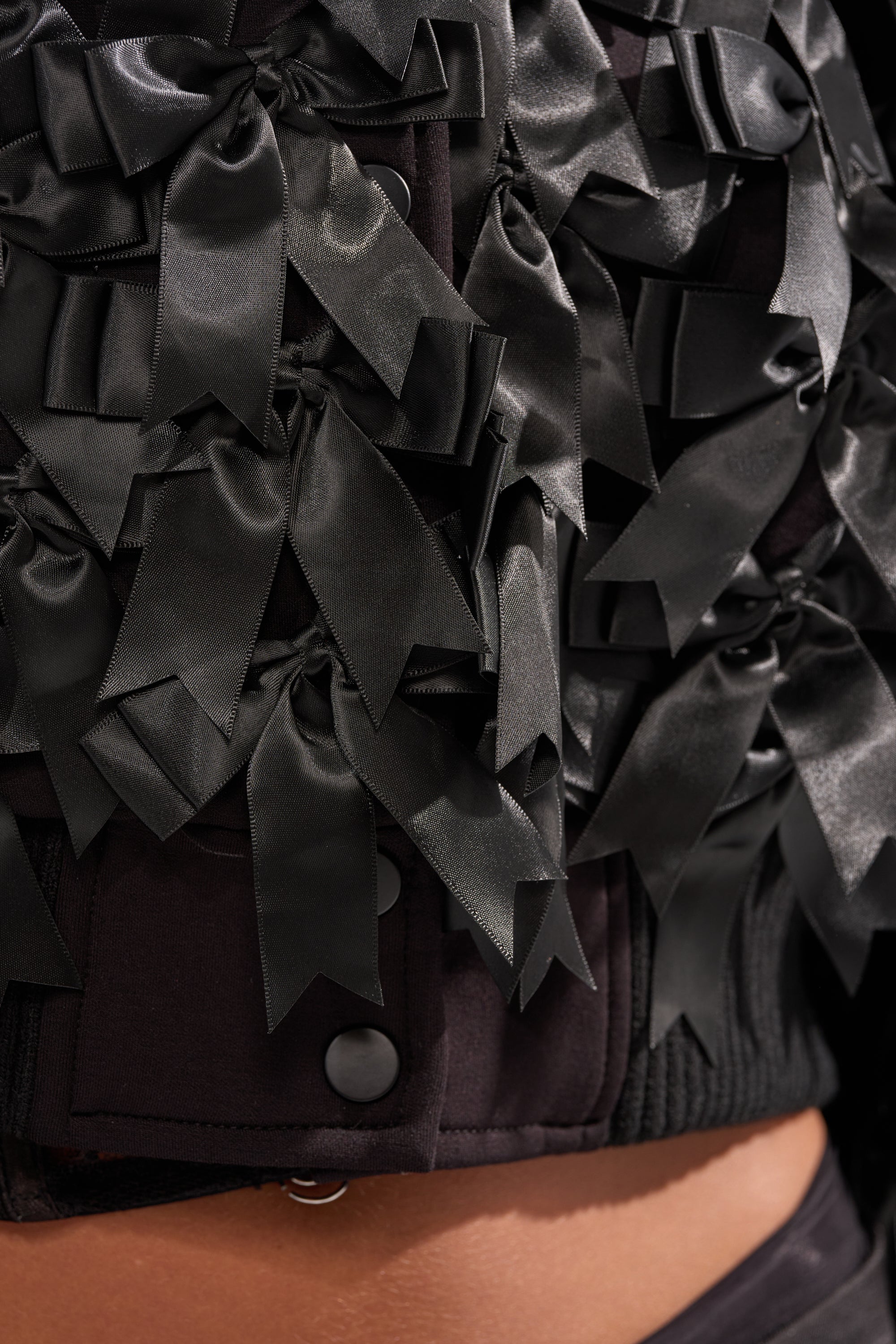 Close-up of the BOW DOWN SATIN BOMBER, featuring glossy black satin bows overlapped for texture, matte black buttons, and a hint of bare skin for a chic finish. 8