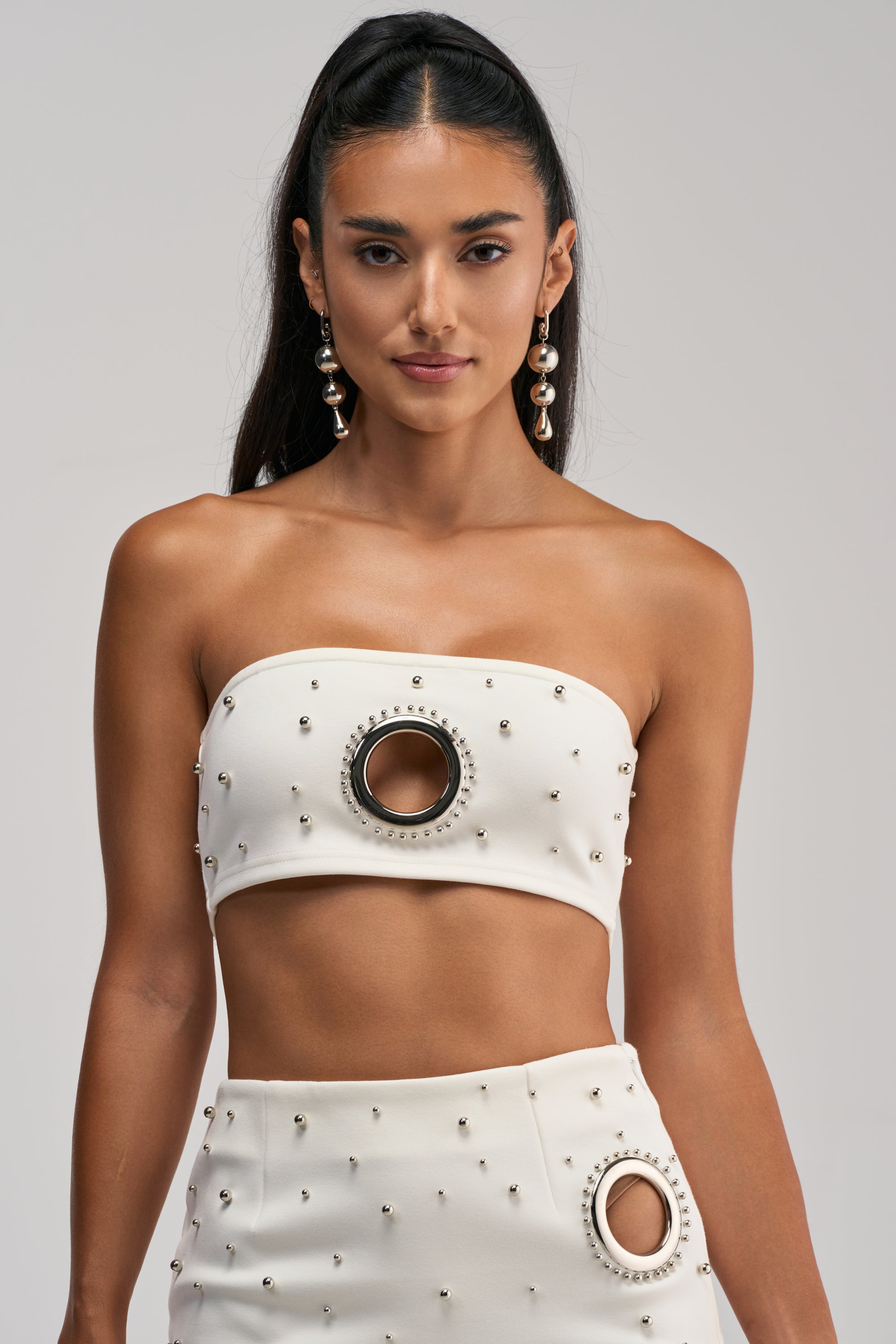 A woman is wearing the SHOW STOPPER BANDEAU IN WHITE, a white beaded strapless crop top featuring a large circular cutout at the center and silver grommet detailing. She pairs it with a matching skirt that has a side cutout and accessorizes with long earrings. Her long, dark, straight hair is styled in a high ponytail.