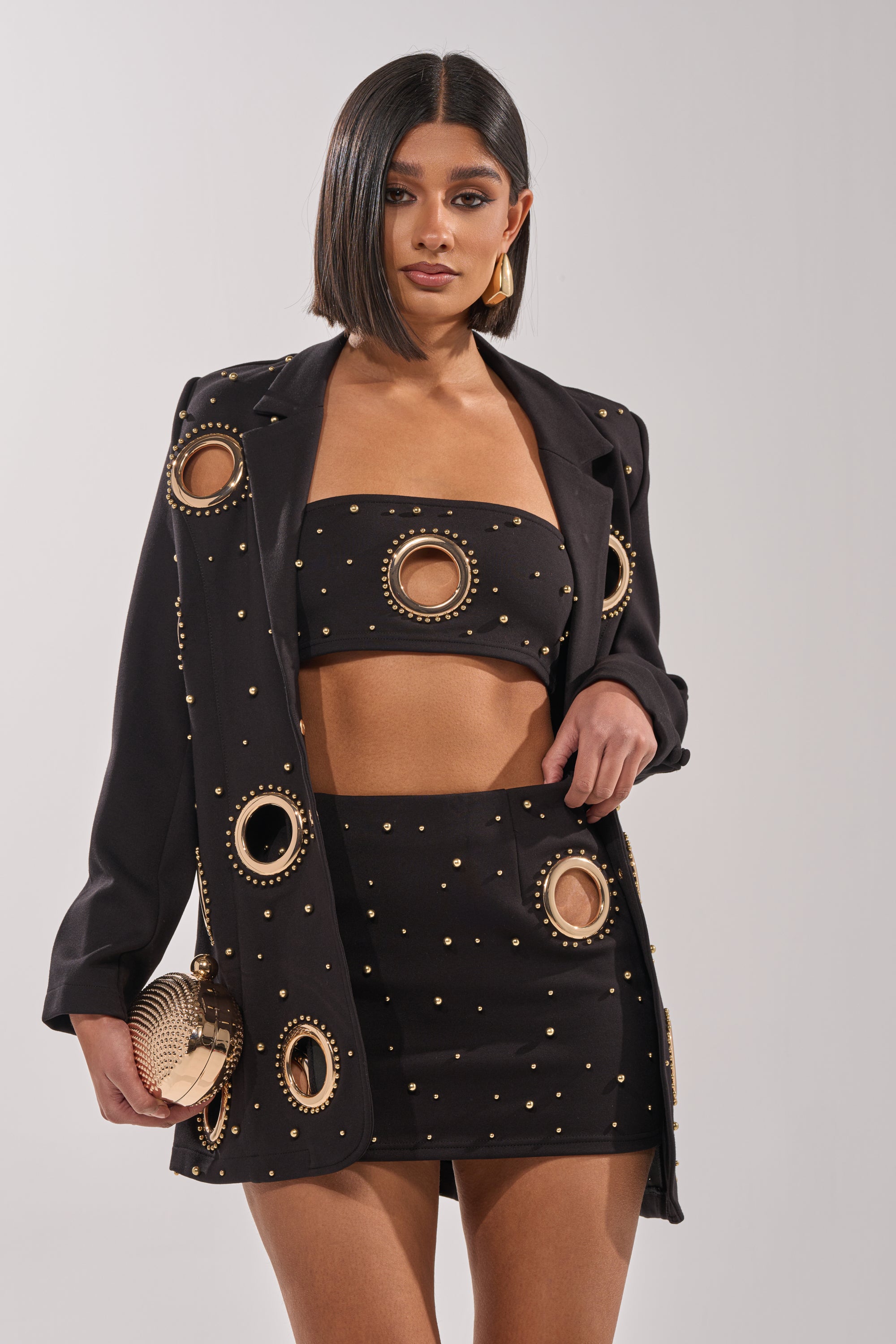 A woman wears the SHOW STOPPER BANDEAU with a matching blazer and mini skirt, all featuring gold-tone studs and large circular accents. She carries a round gold purse and completes her look with geometric earrings. 1