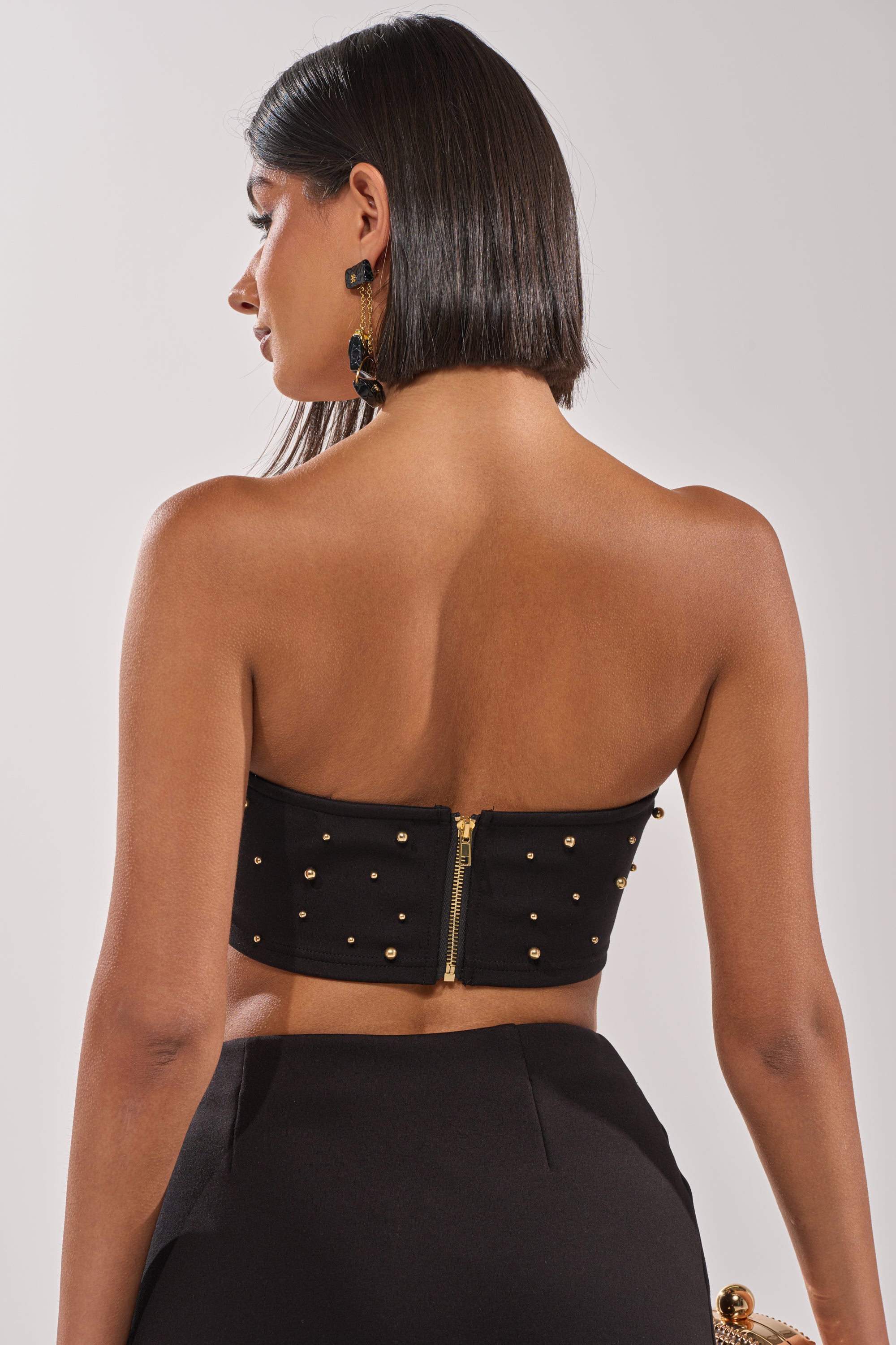 A woman with straight, shoulder-length dark hair is shown from the back wearing the SHOW STOPPER BANDEAU—black with gold studs and a visible zipper—paired with high-waisted black pants. 4