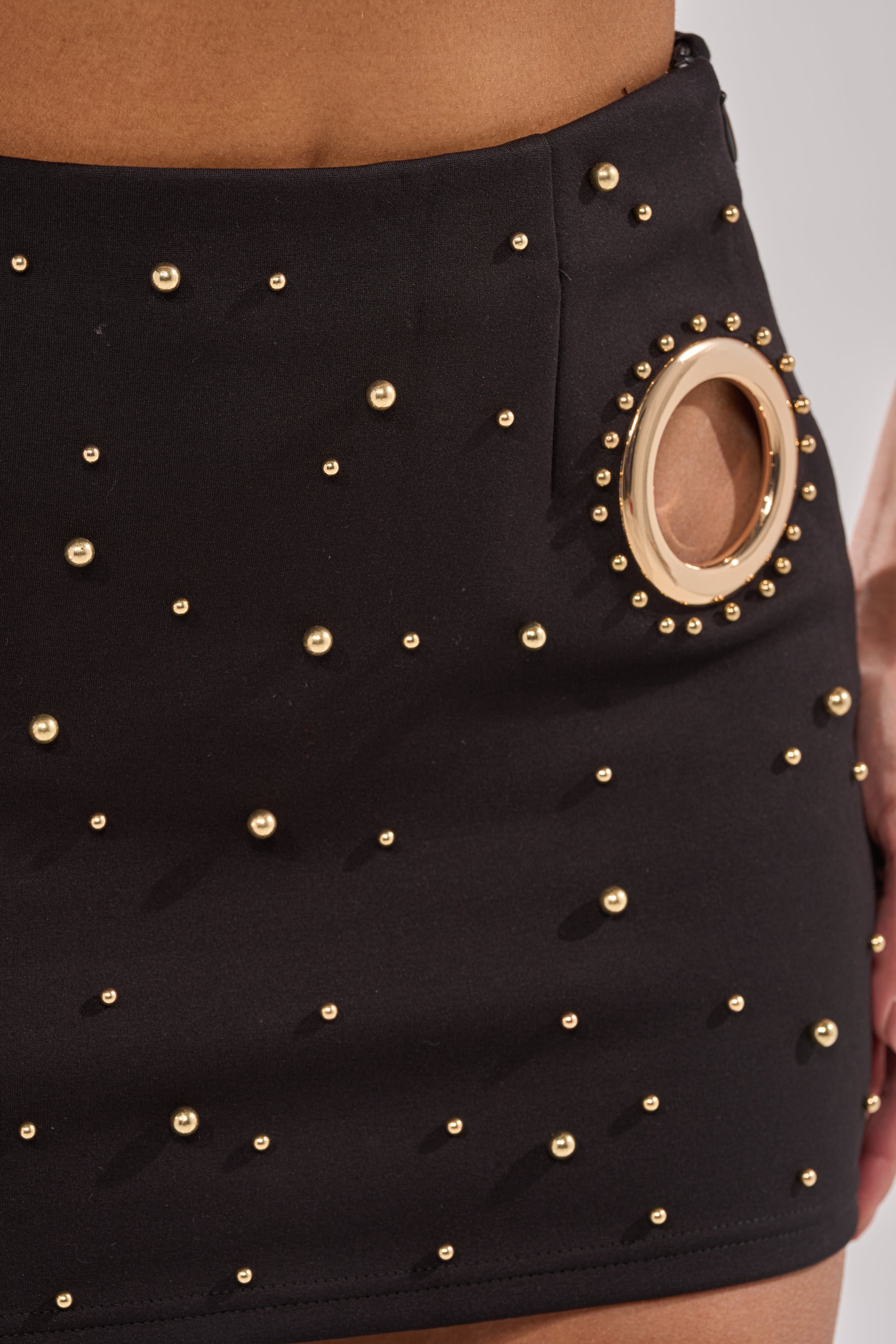 Close-up of the SHOW STOPPER MINI SKIRT, a high-rise black mini skirt featuring small gold studs and a large circular gold grommet detail near the hip. 8