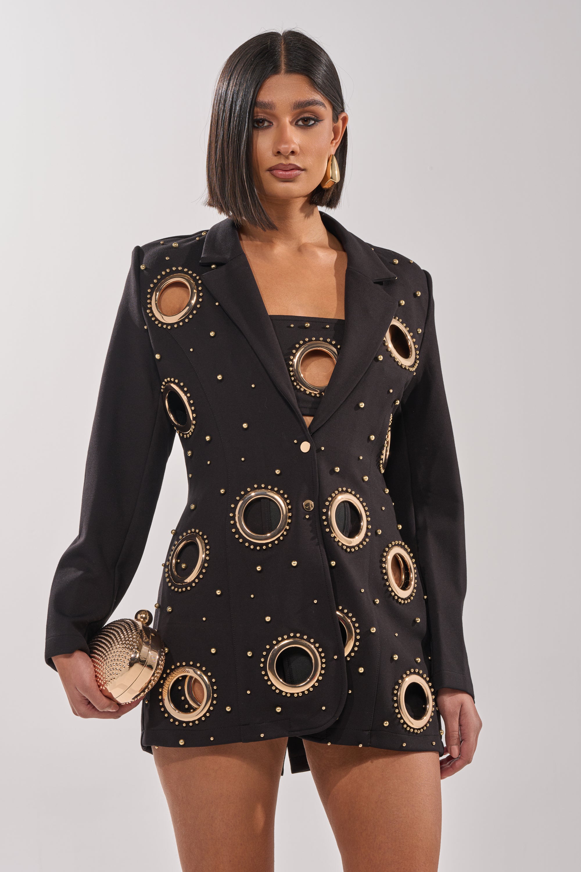 A woman with a sleek bob wears the SHOW STOPPER BLAZER, featuring bold circular gold-outlined cutouts and dome studs. She accessorizes with a textured gold clutch and statement earrings, posing confidently against a plain background. 0